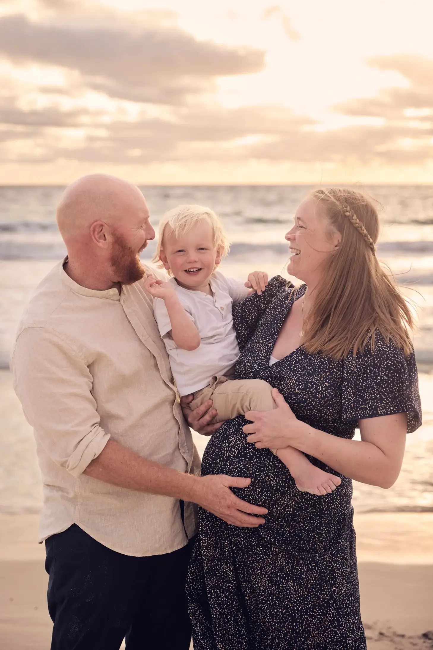 A man and woman are holding a young child at the beach during sunset, all smiling and looking at each other.