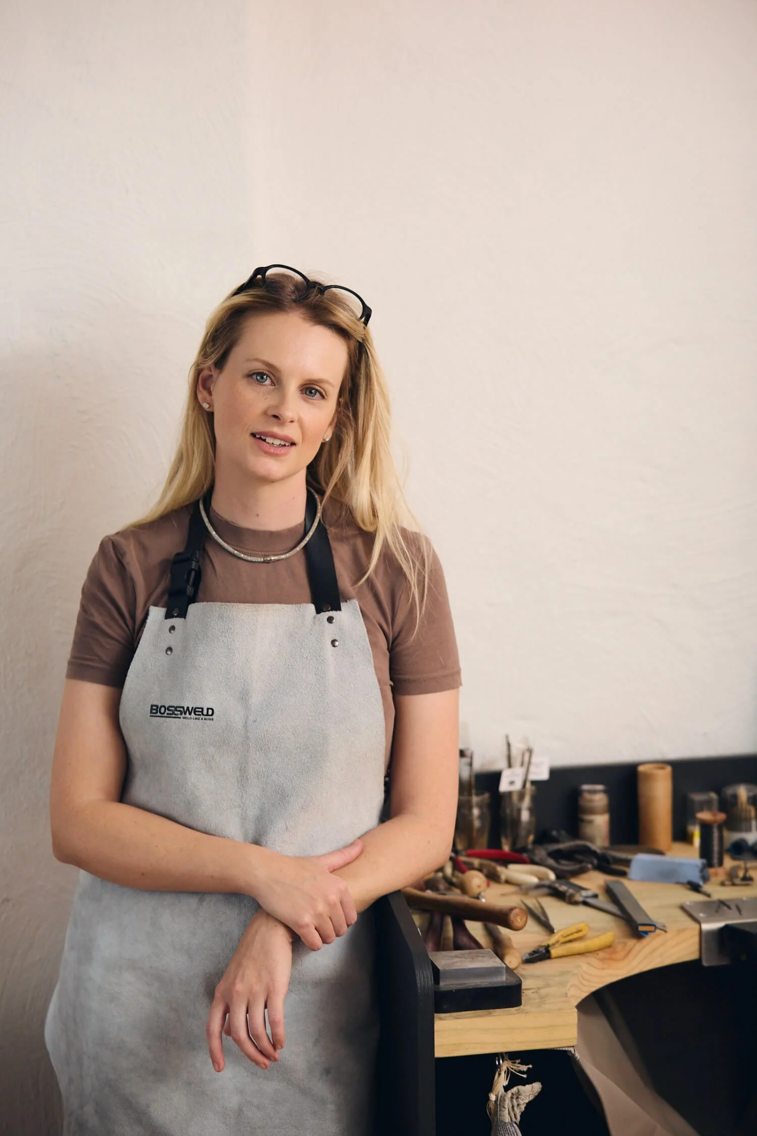 A woman with blonde hair and glasses on her head wearing a beige shirt and a white apron stands in front of a workbench filled with tools and materials in a workshop setting.