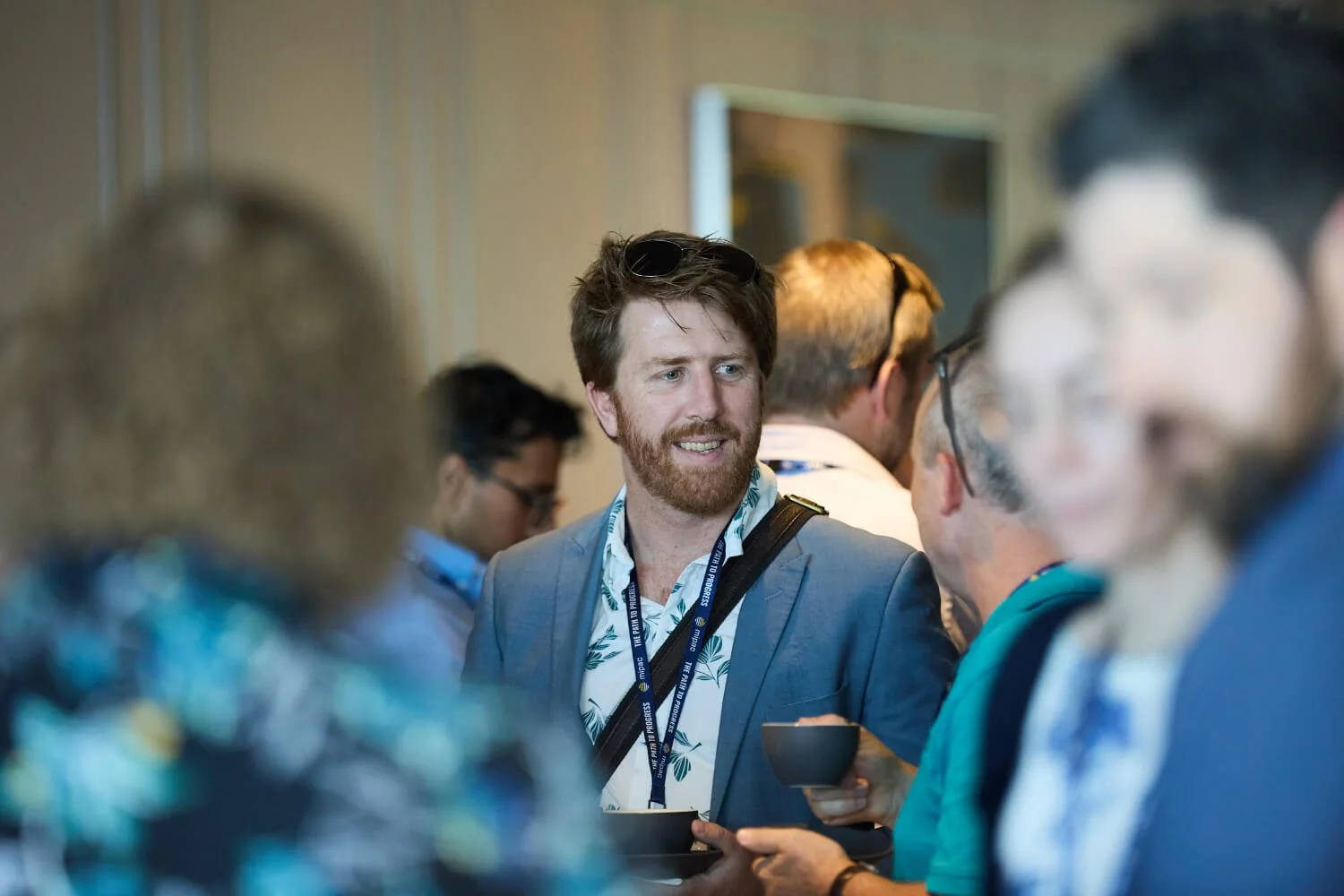 A man with brown hair, beard, sunglasses on his head, wearing a blue blazer and a patterned shirt, is smiling and engaged in conversation at a networking event, surrounded by other attendees.