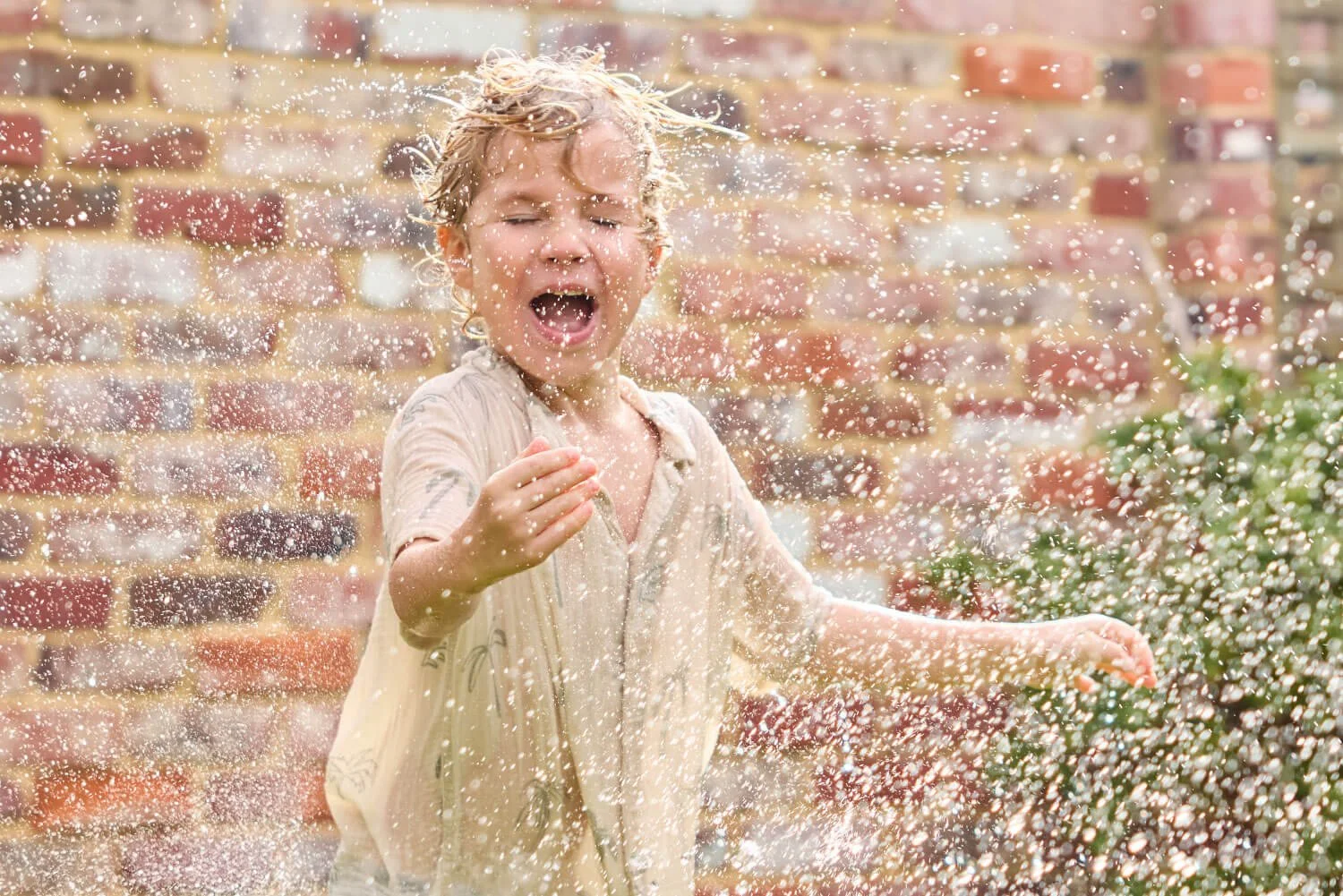 A young boy playing with water outdoors, splashing water with joyful expression, in front of a brick wall.