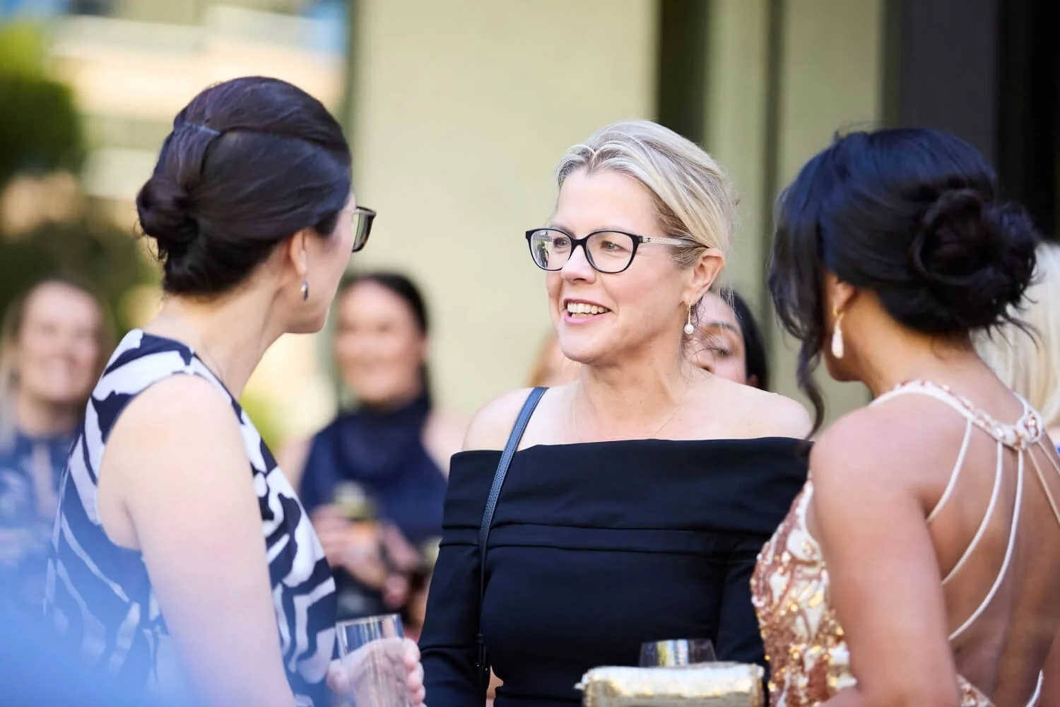 Group of women at social gathering, engaged in conversation, outdoors, dressed stylishly, holding drinks.