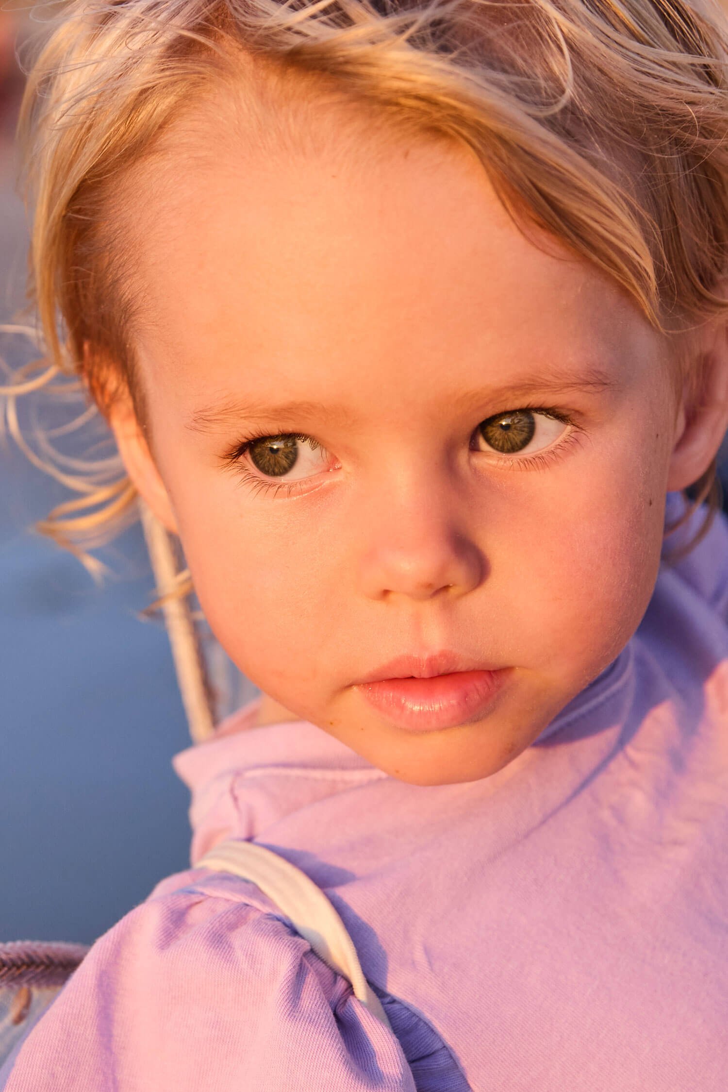 Close-up of a young child's face with curly blonde hair and a pink shirt, outdoors in natural light.