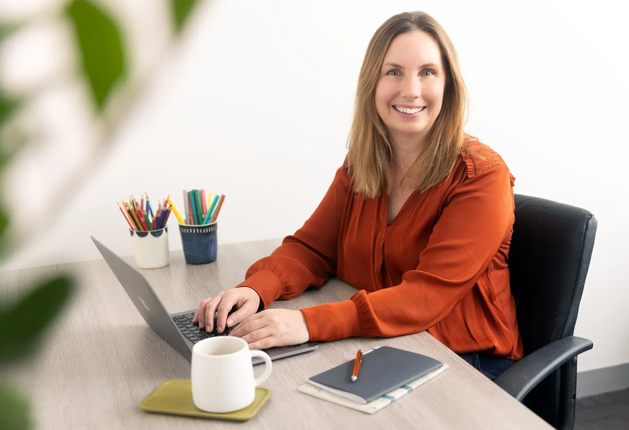 A woman with light brown hair is smiling while typing on a laptop. On the desk are a mug and a notebook and two cups of coloured pencils in the background.