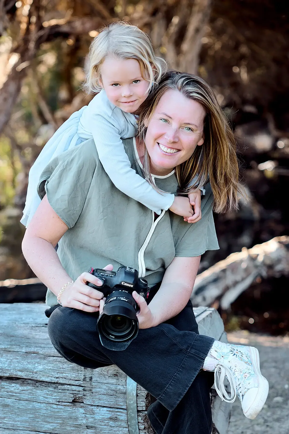 A woman sitting outdoors on a wooden log with a camera in her hands, and a young girl with blonde hair on her back, both smiling. The background is blurred with trees and natural scenery.