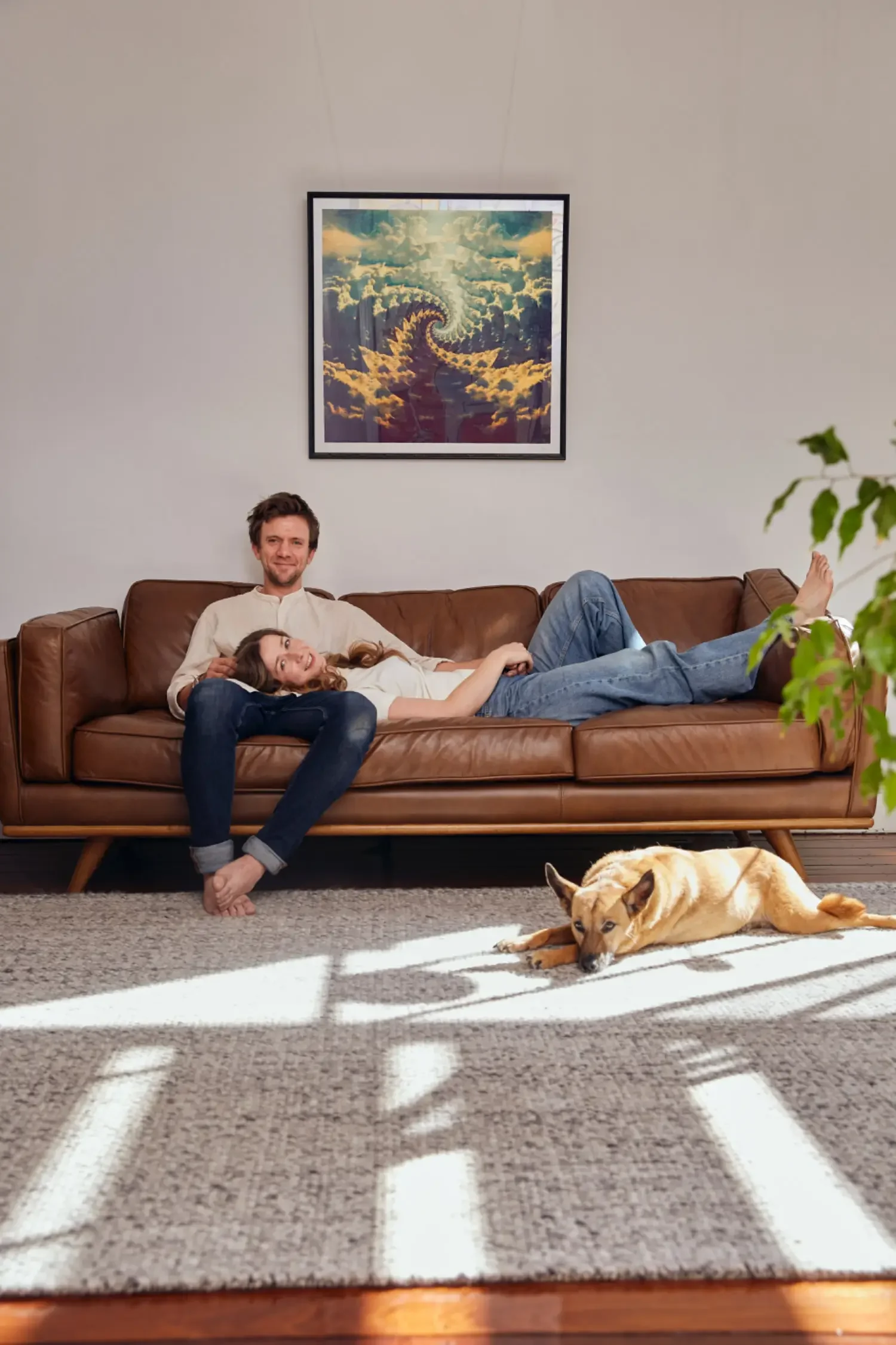 A man and a woman are relaxing on a brown leather couch in a living room. The man is seated and smiling, the woman is lying down with her head on his lap. A dog is laying on the carpet in front of the couch. The living room has a framed painting of a surreal, spiral staircase in the clouds hanging on the wall and a green plant in the corner. Sunlight is streaming through blinds, casting shadows on the carpet.