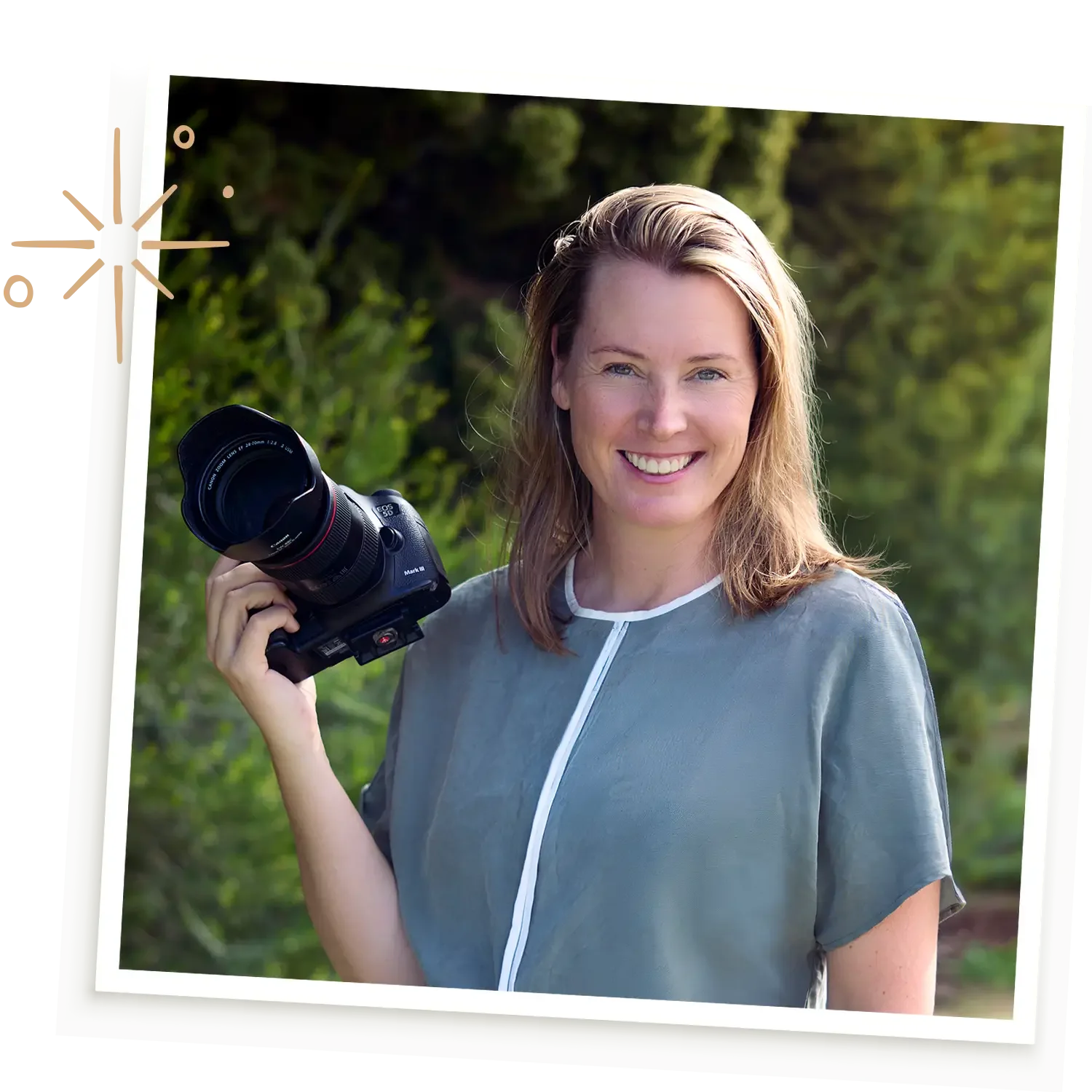 A woman outdoors holding a camera, smiling, with trees in the background.