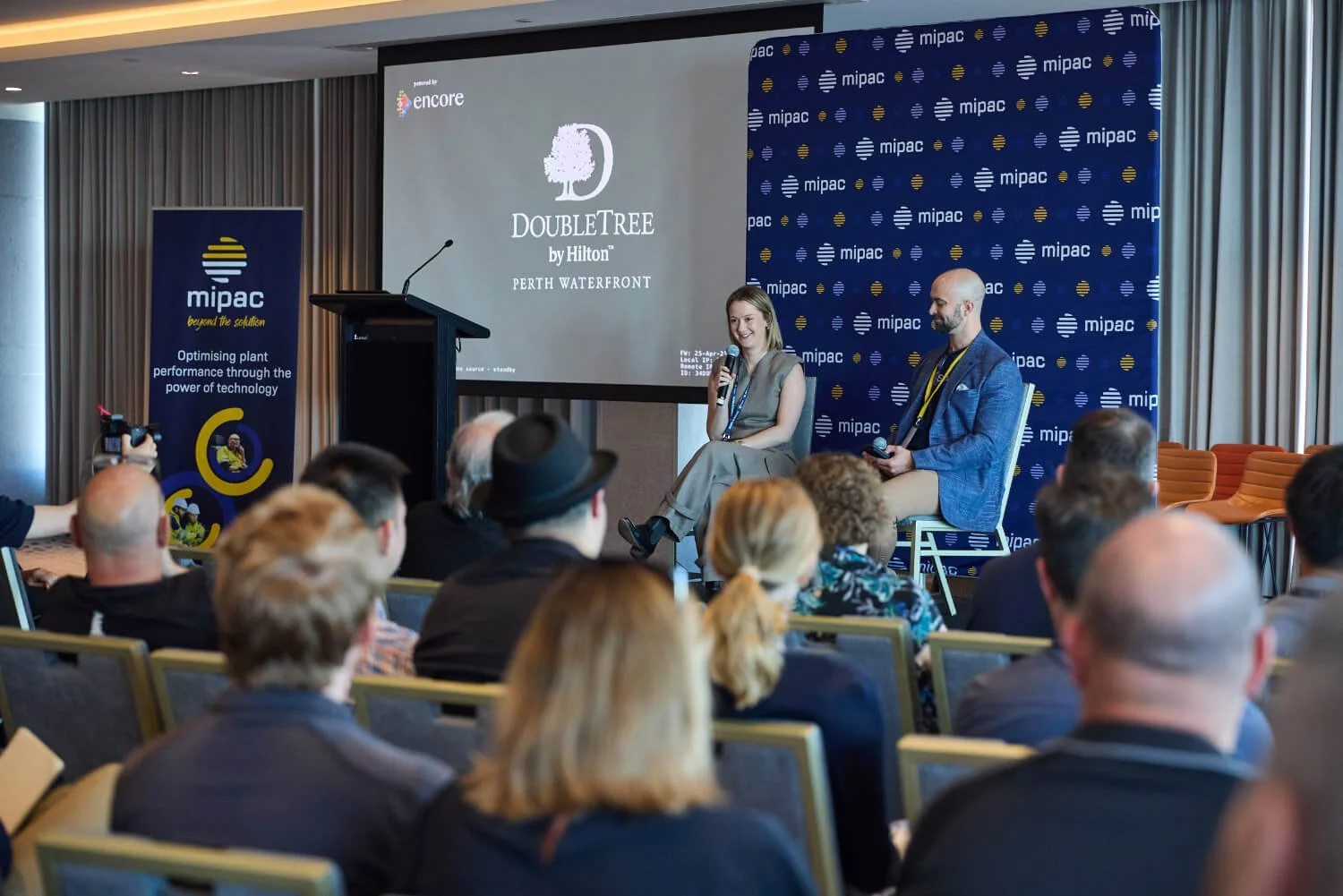 A woman and a man seated on stage during a conference, with an audience watching. The stage has a large screen displaying the 'DoubleTree by Hilton Perth Waterfront' logo, and a MIPAC banner is visible on the left side. The woman is holding a microphone, and both are wearing conference badges.