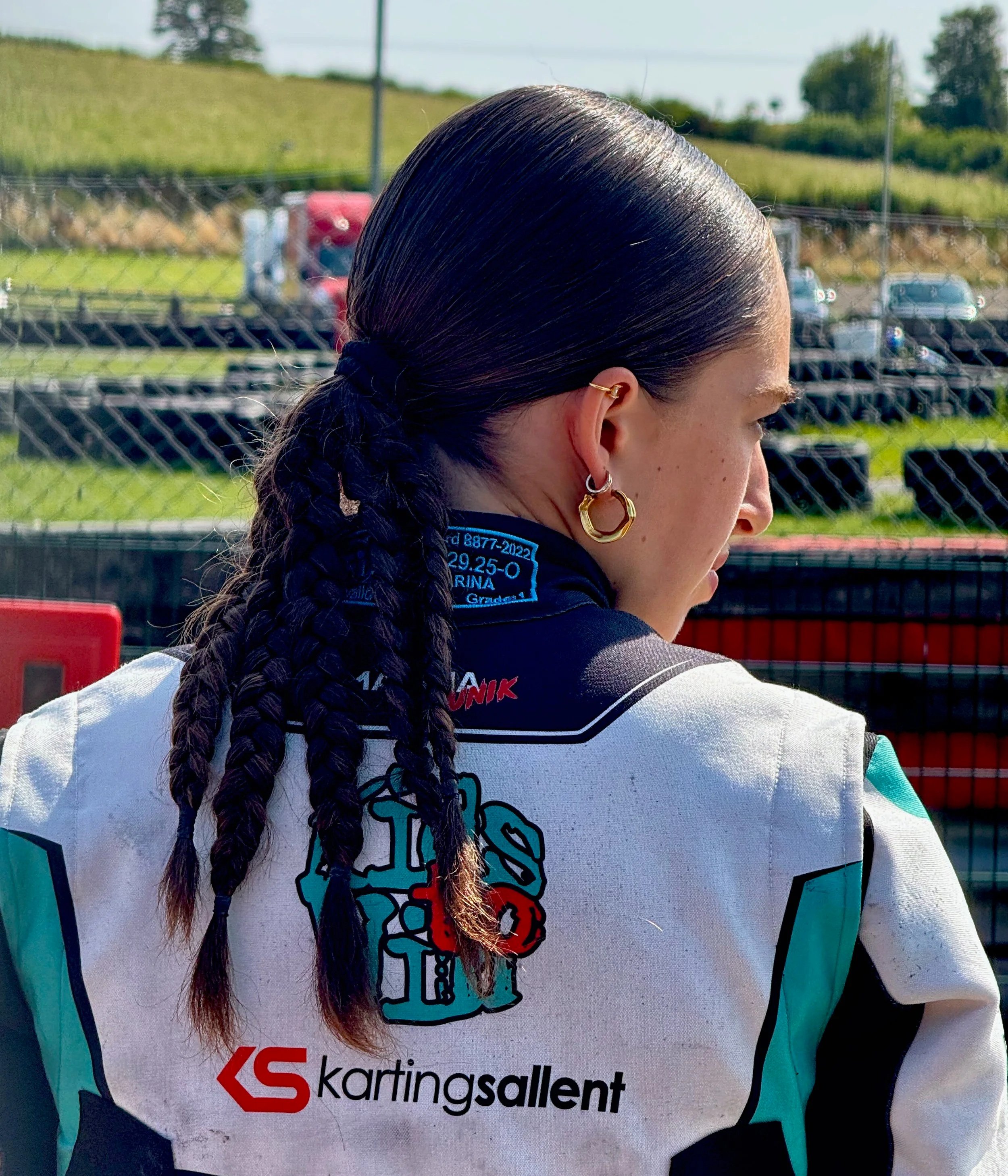 Close-up of a woman with long, dark hair tied in a braid, wearing racing gear with a logo for 'kartingsallent' and a graphic of a skull with a helmet, outdoors with a chain-link fence and vehicles in the background.