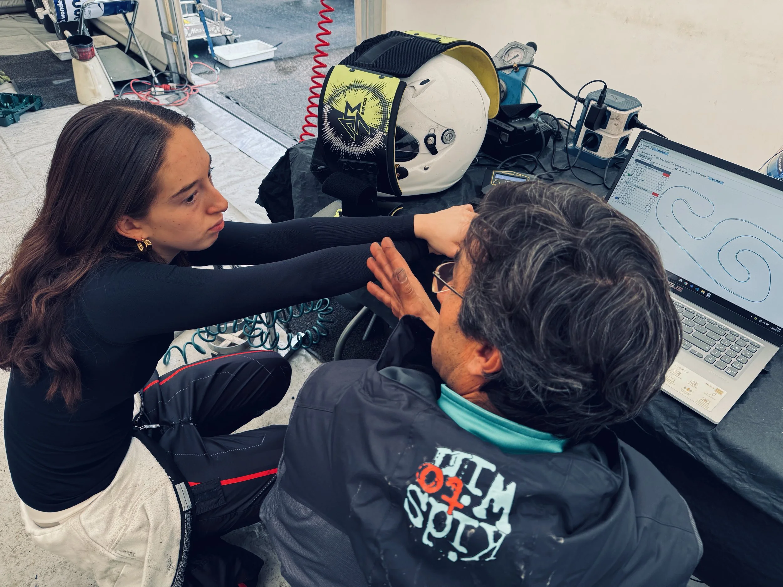 Two individuals, one young woman and one older man, sitting at a table with a computer monitor, a racing helmet, and electronic equipment, appearing to analyze data or racing track information.