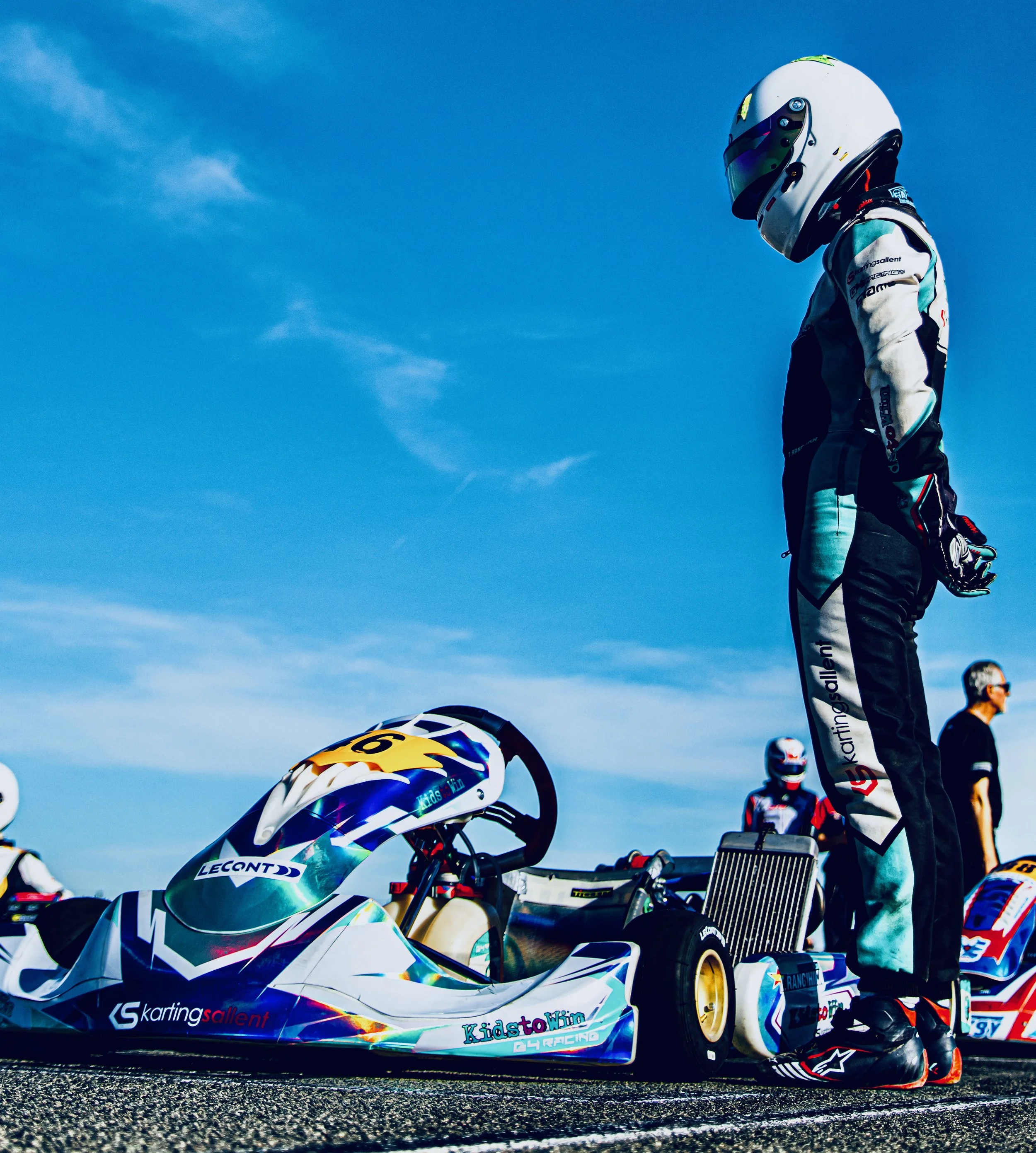 A race car on a track with a helmet and racing gear, and a person standing in the background during daytime.