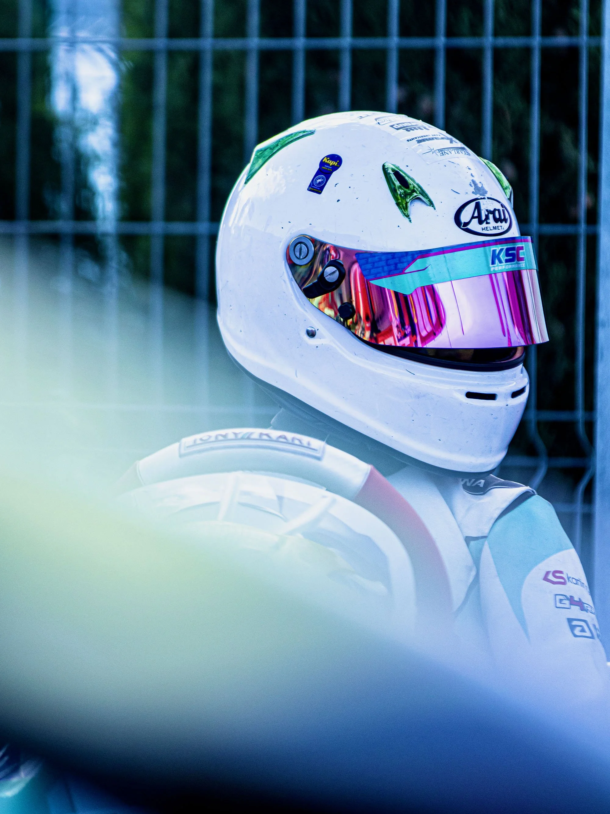 Close-up of a white racing helmet with black, purple, and blue accents, resting on a reflective surface with a blurred background of a metal fence and trees.