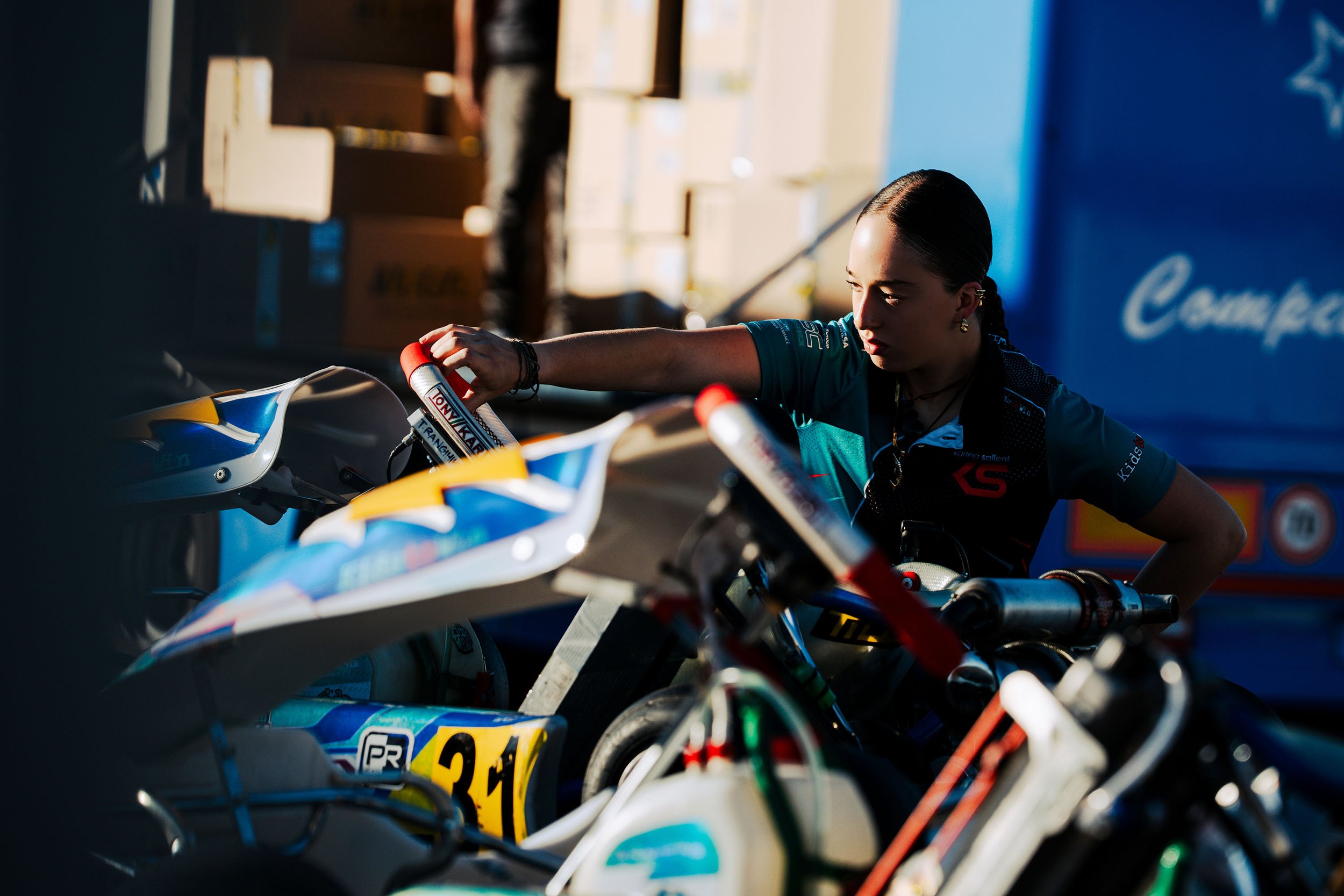 A young woman with dark hair tied back, wearing a blue racing jersey, is getting ready on her motorcycle at a race track. There are other motorcycles and a blue sign with white text in the background.