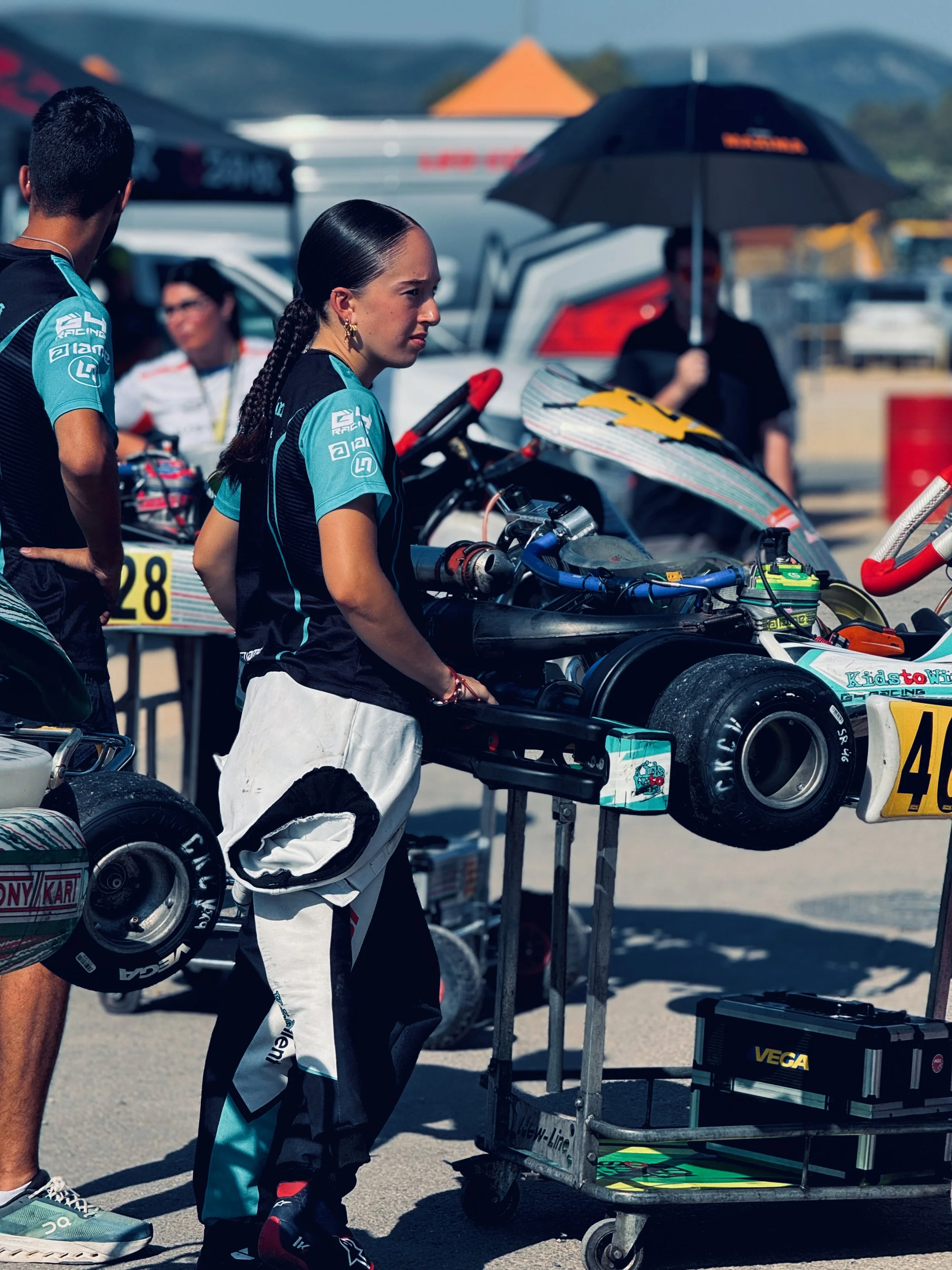 A woman in racing gear standing at a go-kart maintenance station with a racing helmet and a toolbox nearby. Other people and go-karts are visible in the background at a racing track.