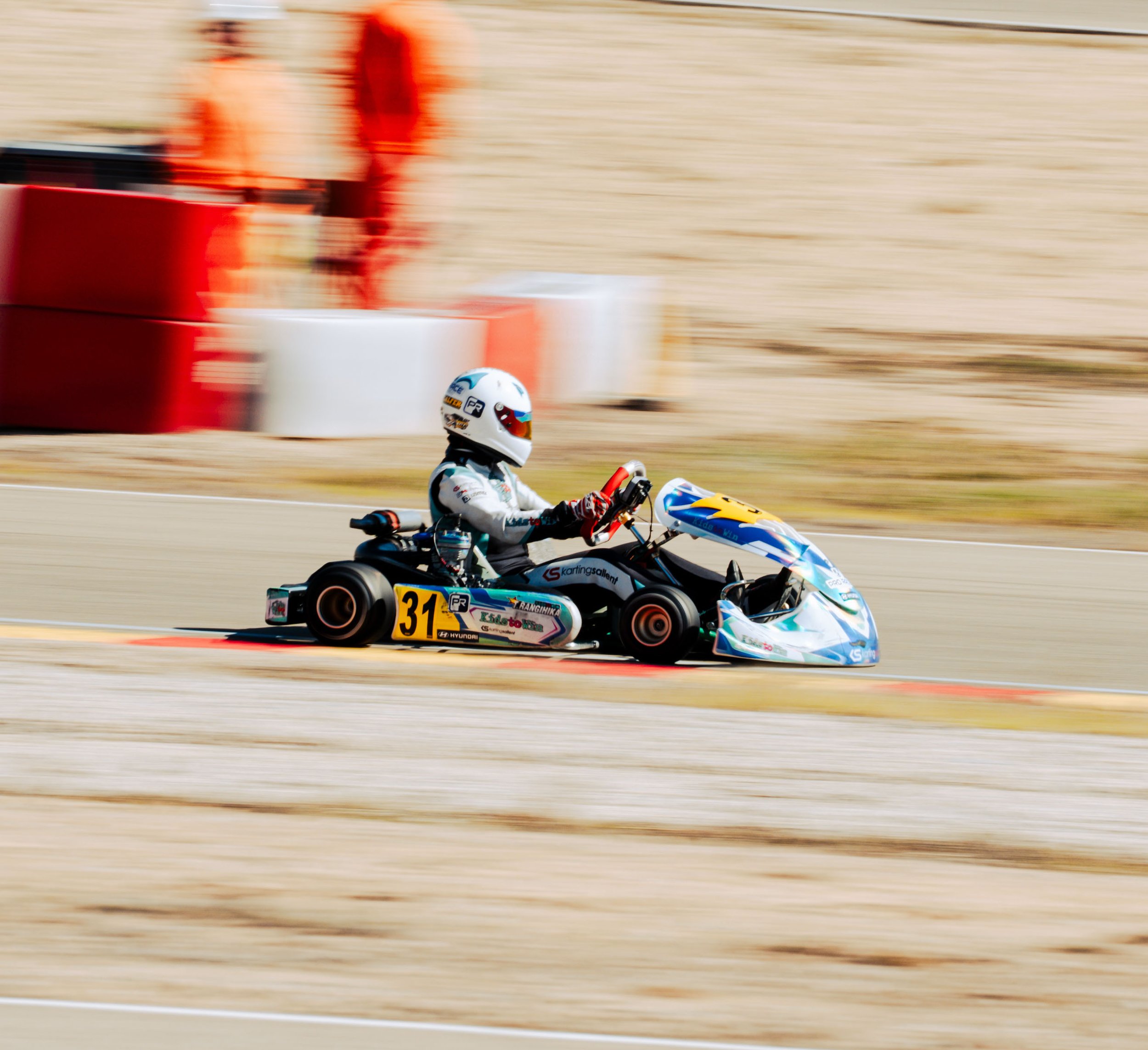A go-kart race with a driver wearing a white helmet and racing suit, speeding on a track with red and white barriers and blurred background.