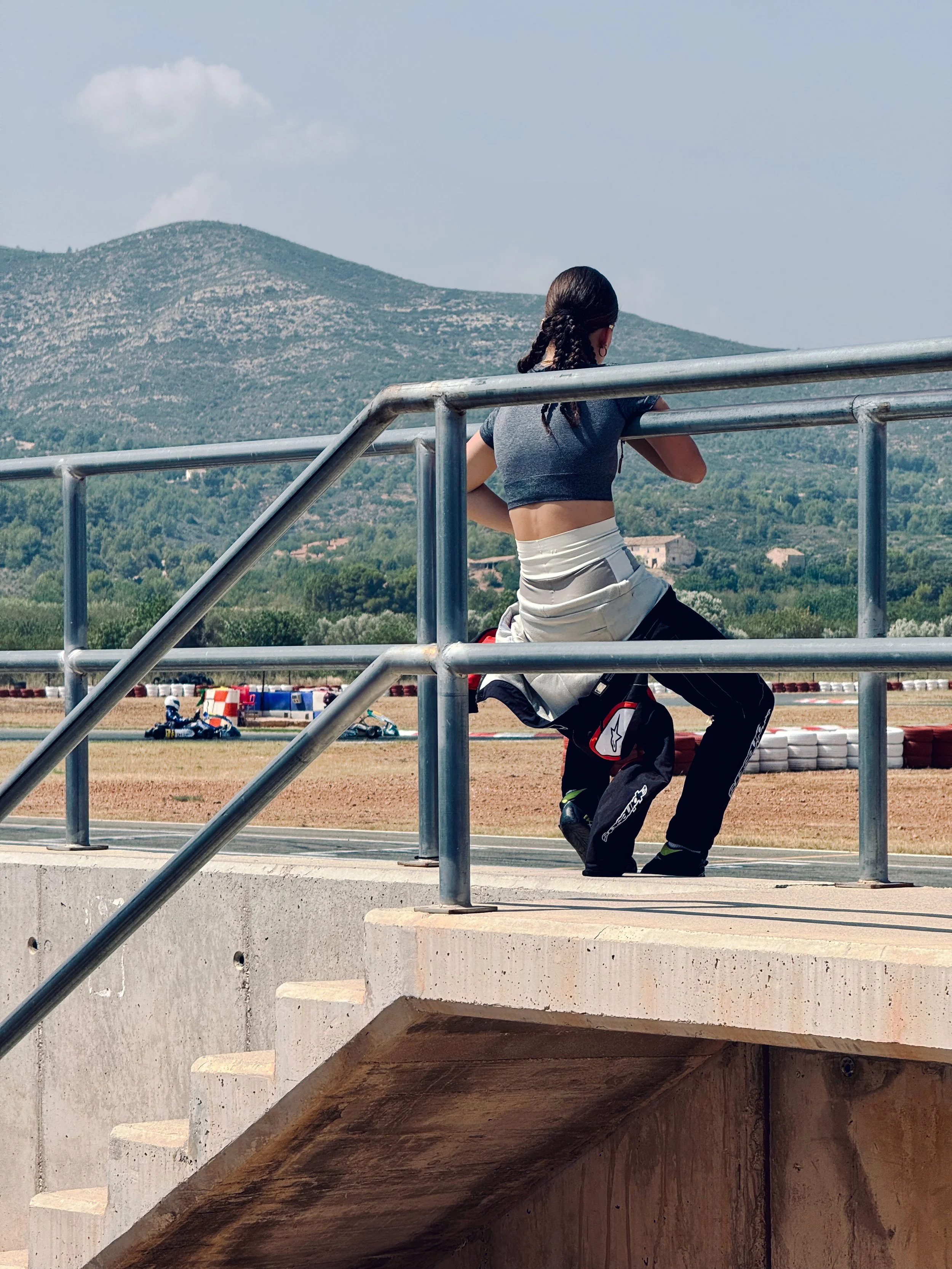A person with braided hair is kneeling on the ground near a stairway with metal railing, looking at a racing track with go-karts in the background and mountains in the distance.