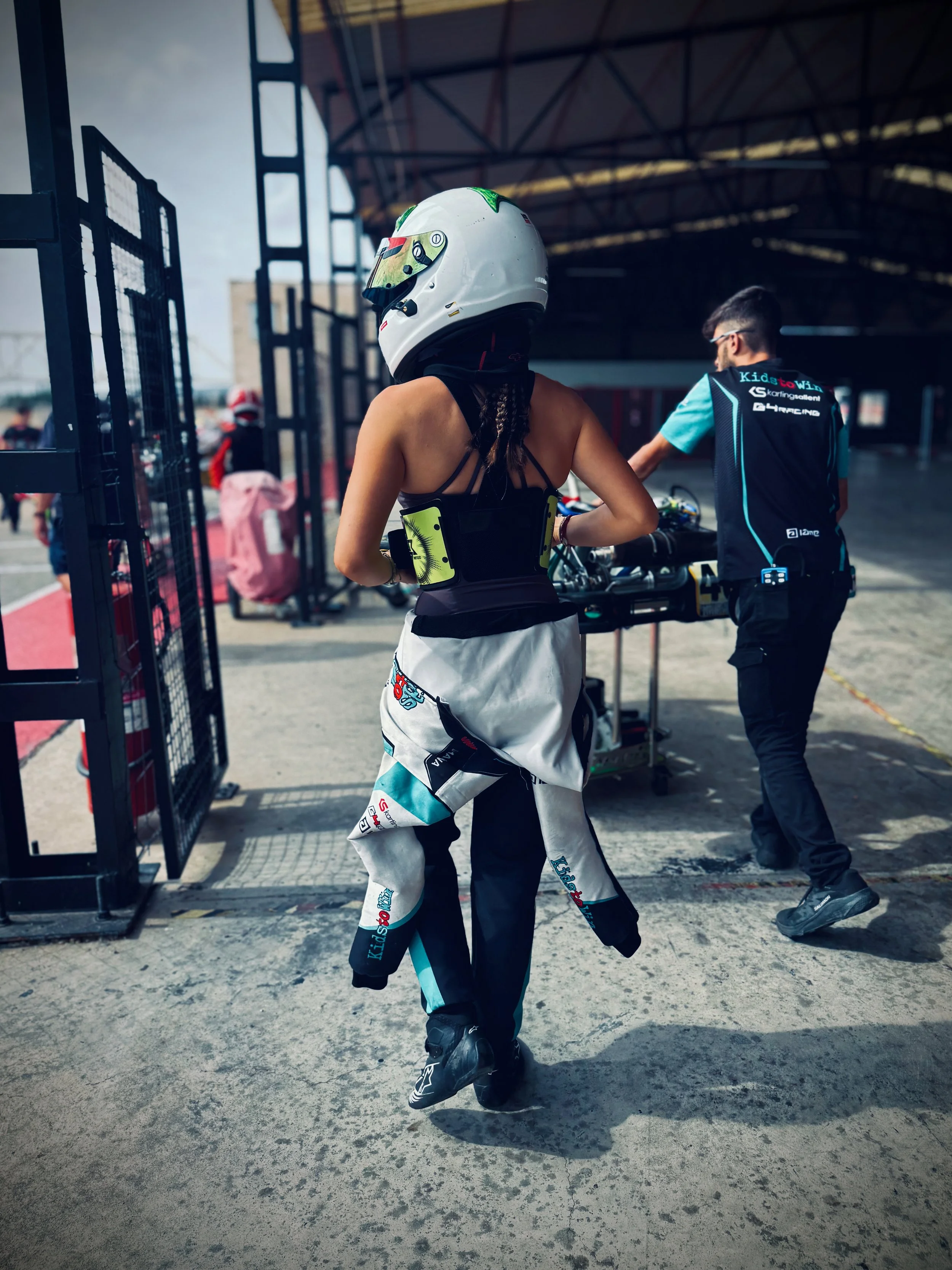 A woman wearing a racing helmet, racing suit, and racing shoes appears to be preparing for a race or practice session at a racing circuit, with pit lane activity visible in the background.
