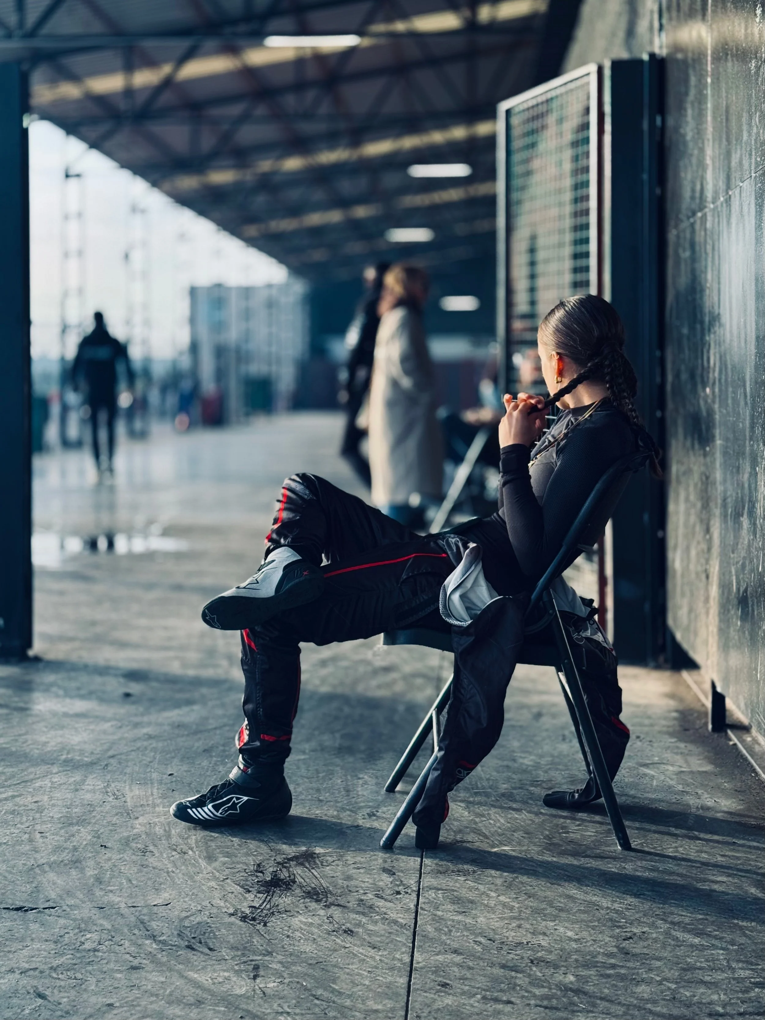 A young woman with braided hair sitting on a chair in a sports or motorcycle gear, holding a riding helmet, in a shaded outdoor setting with a reflective surface on the wall behind her. Blurred figures walking in the background.