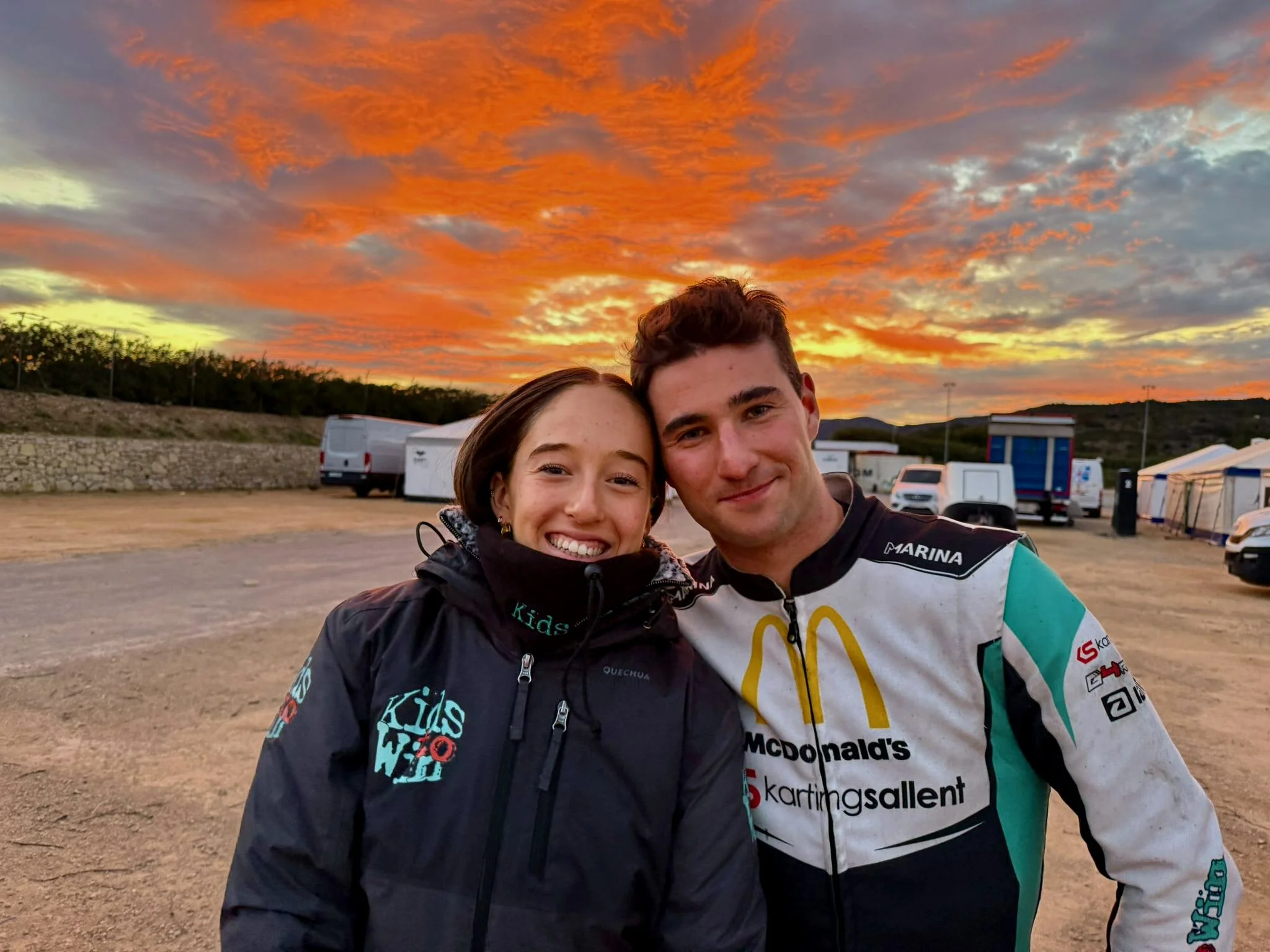 Two young people, a woman and a man, smiling and posing for a photo outdoors during sunset. The woman has long brown hair and is wearing a dark jacket, and the man has short brown hair and is wearing a racing sports shirt with logos, including McDonald's and karting sallen. The background features a colorful, cloudy sky with hues of orange, pink, and purple, along with parked vehicles and ten