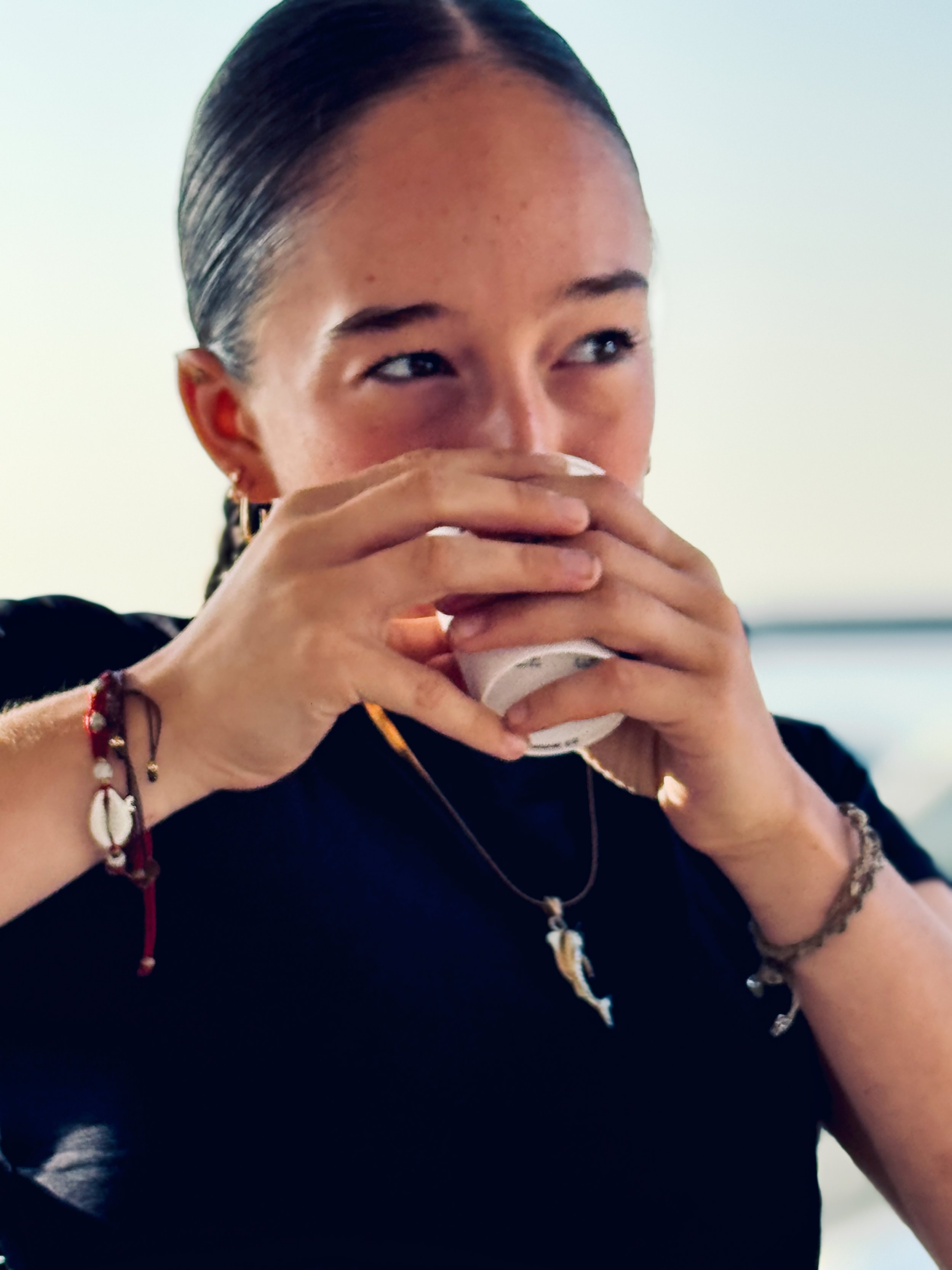 A young woman with dark hair pulled back, covering her mouth with her hands, wearing multiple bracelets and necklaces, in a casual indoor setting.
