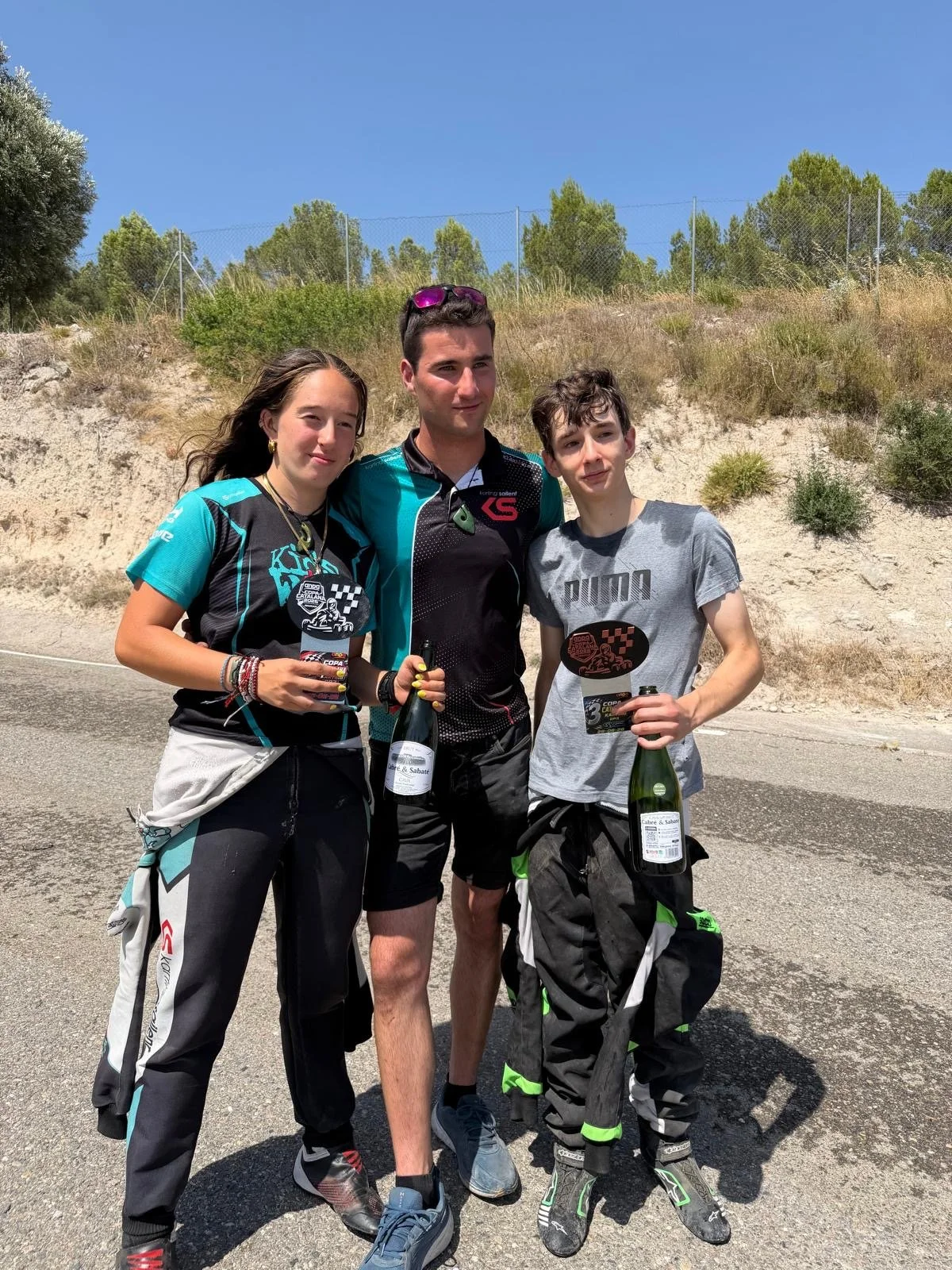 Three young people standing outdoors on a sunny day, holding trophies and bottles of champagne, celebrating a sports event. Two boys and one girl, dressed in casual and sportswear, with a background of a dirt hill, bushes, and a clear blue sky.