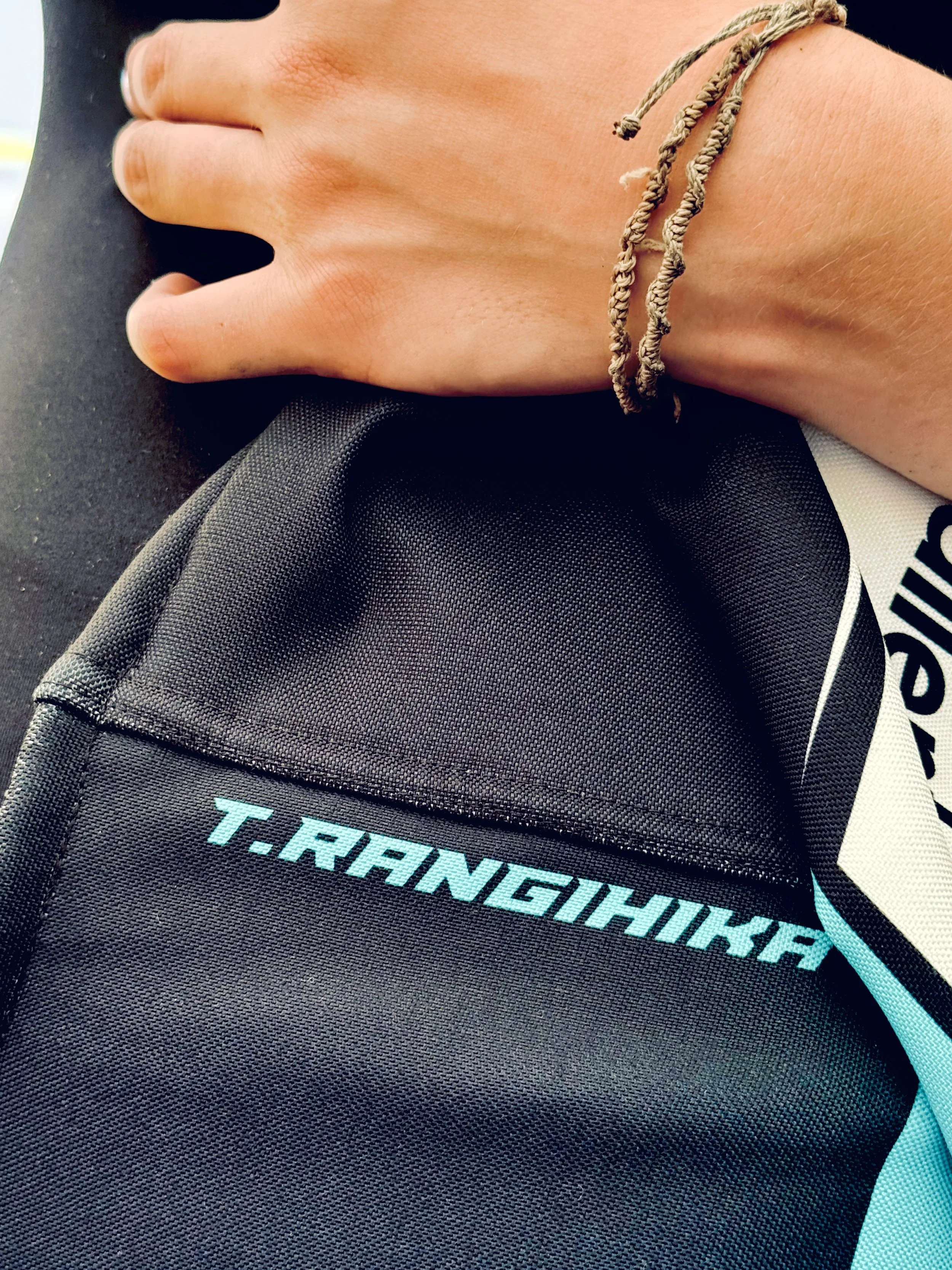Close-up of a hand resting on a black sports bag, showing a beige rubber bracelet on the wrist and part of a shirt with the phrase 'T.RANGEH' in light blue text.