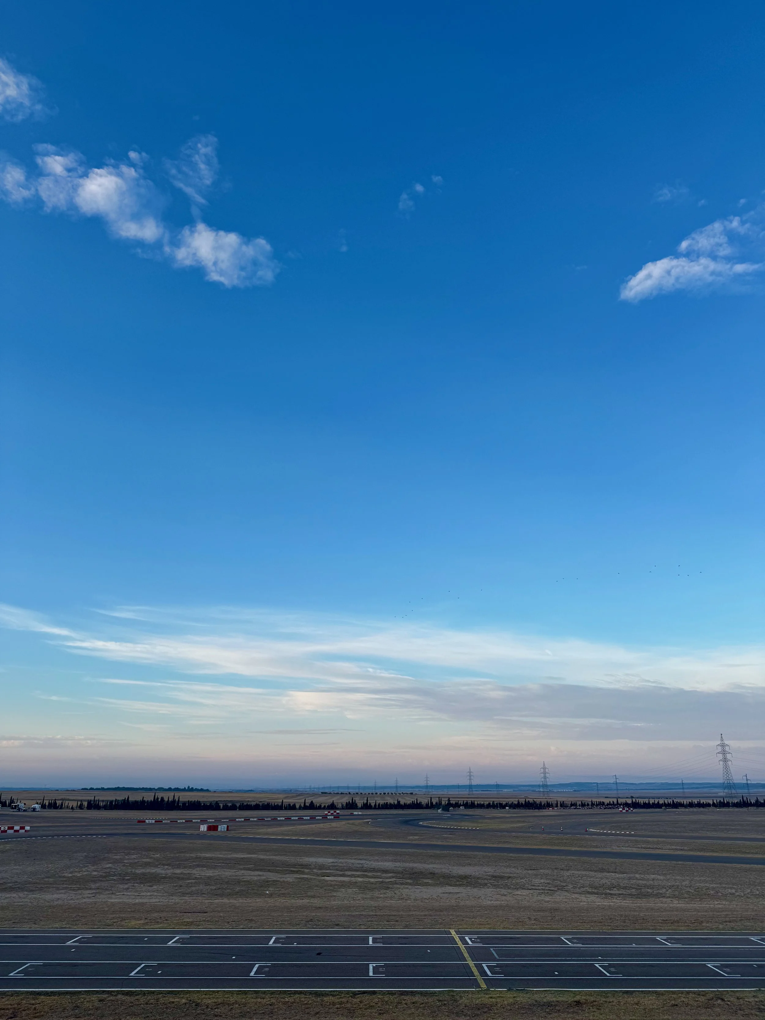 Wide view of a karting track in the foreground, open flat land and distant power lines under a clear blue sky with few clouds.