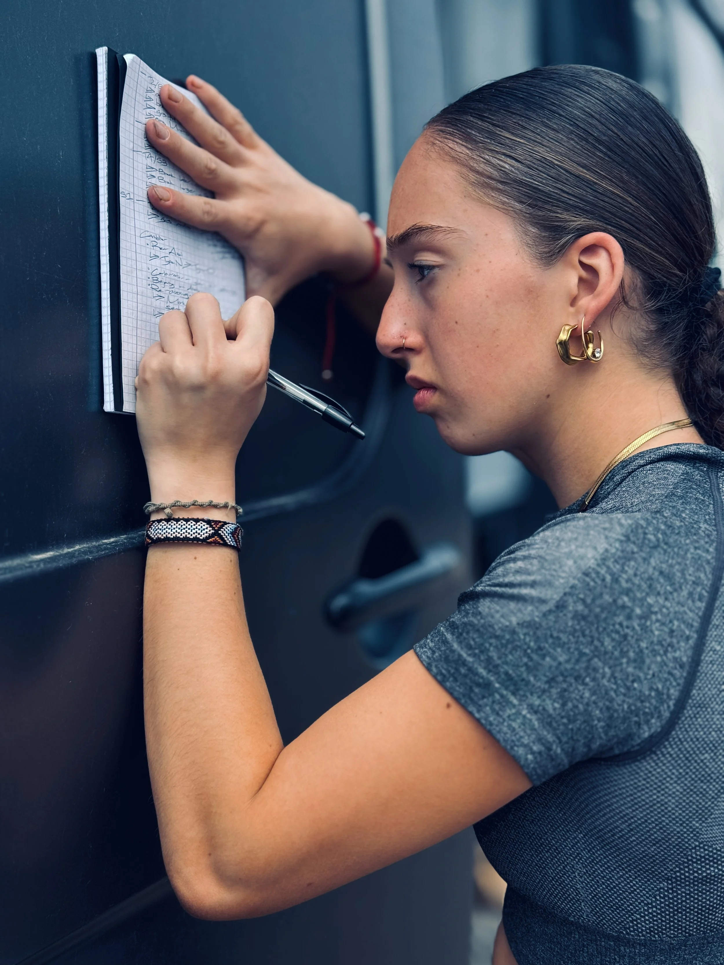 Taya, wearing a gray t-shirt, gold jewelry, and earrings, writing notes on a small spiral notebook pressed against a blue wall.