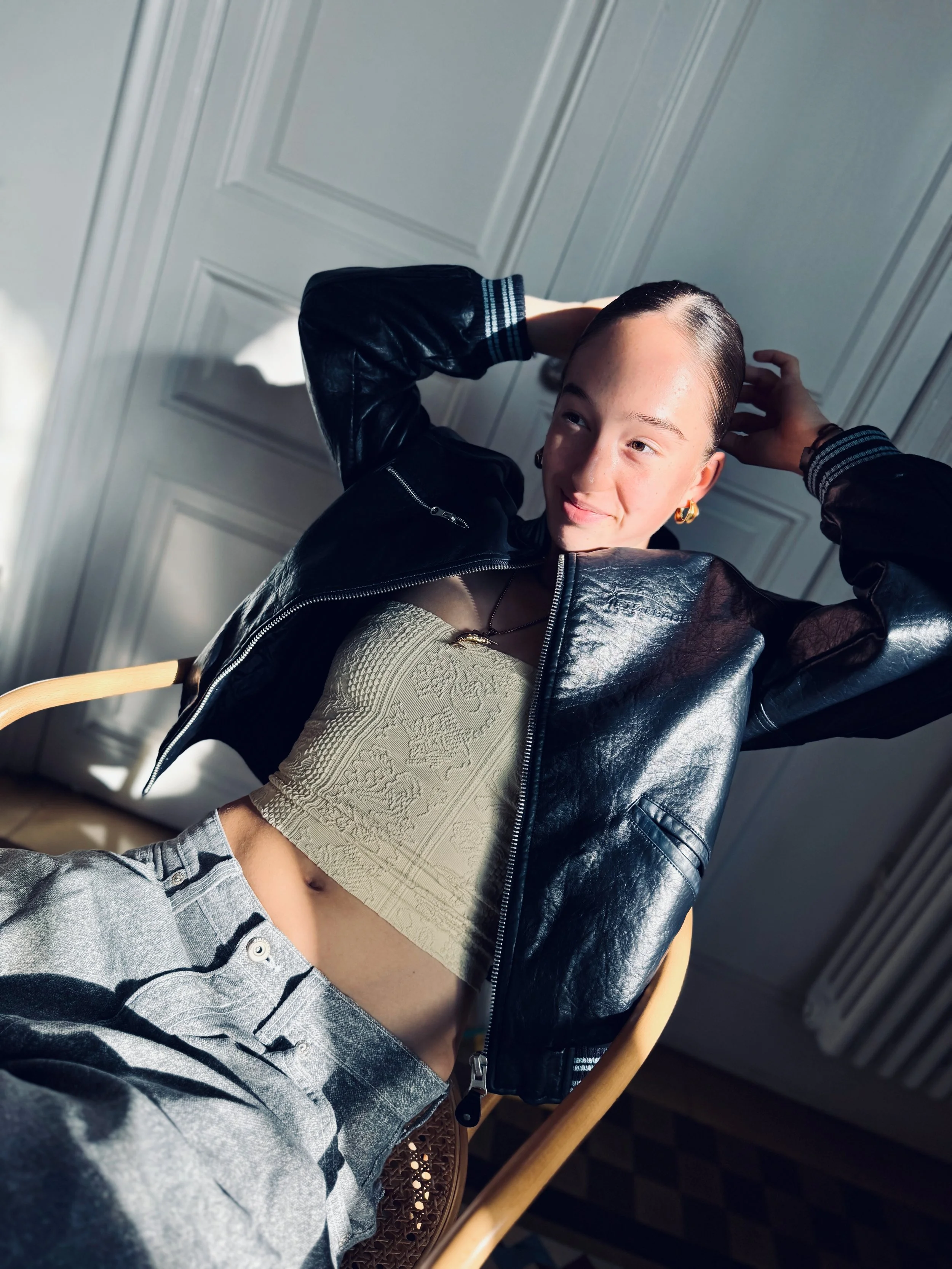 A young person with short dark hair, wearing a black leather jacket, beige top, and gray checkered pants, is sitting in a wooden chair near a white paneled wall. They are smiling and have their arms raised behind their head, with sunlight shining on their face.