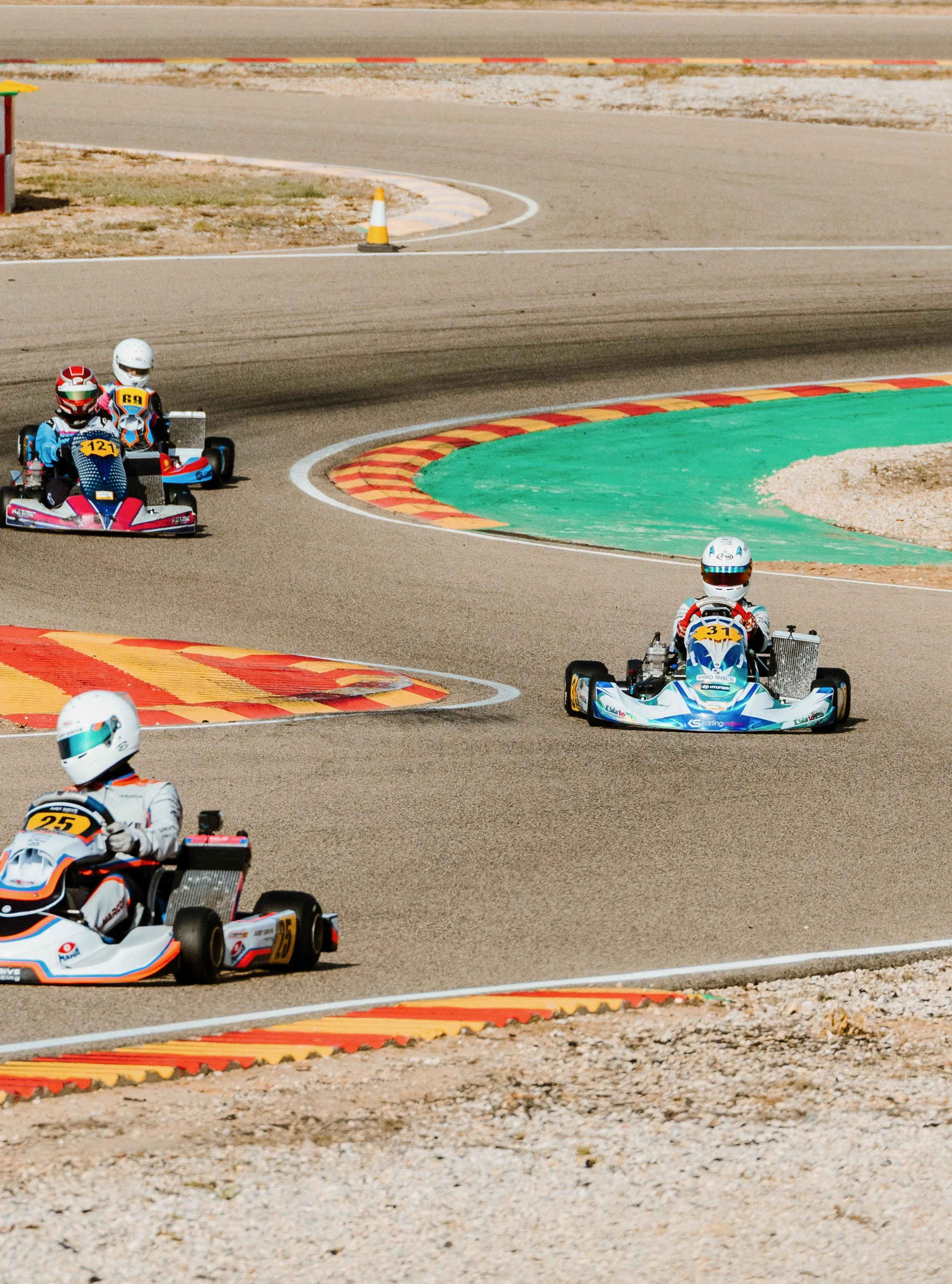 Multiple go-karts racing on a paved track with colorful curbs and orange cones, with drivers wearing helmets.