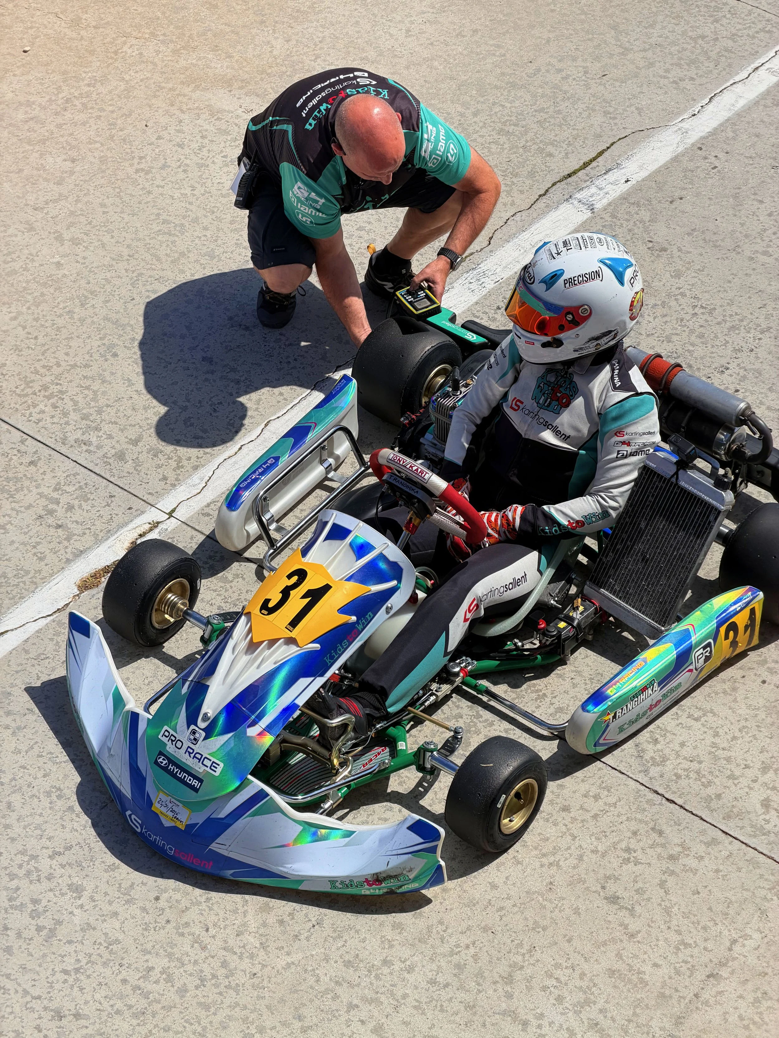A go-kart with the number 31 on it, painted with iridescent colors, is being serviced by a man in a black and teal uniform. The man is bending over, holding a tool, while the driver is seated in the go-kart wearing a white racing suit and a helmet with a blue and orange visor.