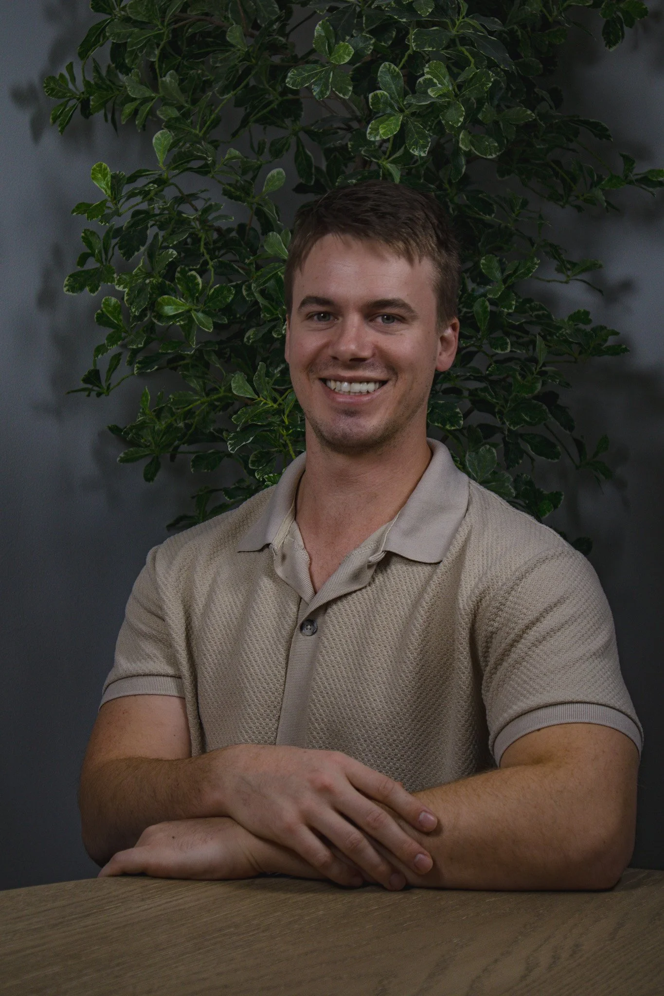 A young man with short brown hair, smiling, sitting at a table with arms crossed. Behind him, there is a leafy green plant against a dark gray wall.
