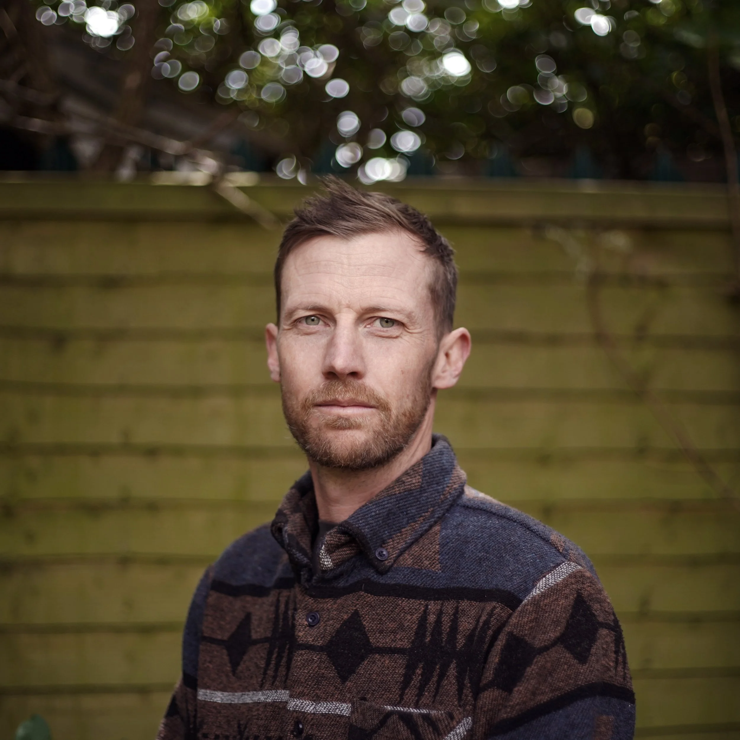 Close-up of a man with short brown hair, light skin, and blue eyes, wearing a patterned brown sweater, standing outdoors in front of a blurred background with wooden fence and leafy trees.