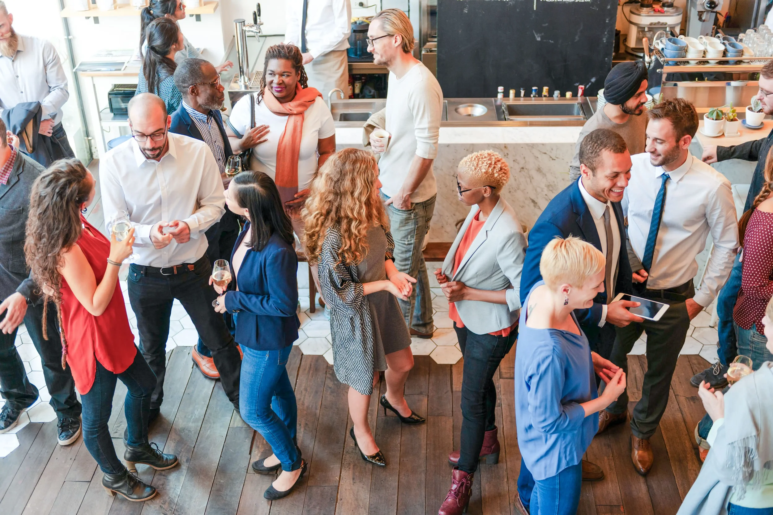 People socializing at a restaurant or cafe, some holding drinks, engaged in conversations, with a bar and kitchen area in the background.