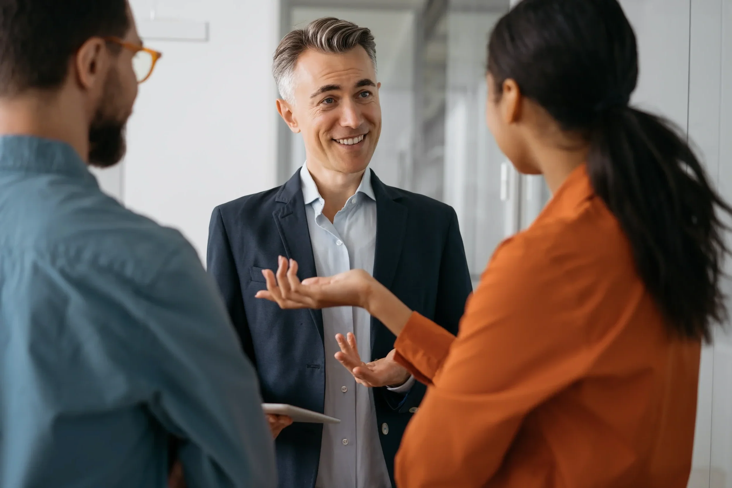 Three people in professional attire having a conversation, with one man smiling and gesturing with his hand, in a modern office setting.