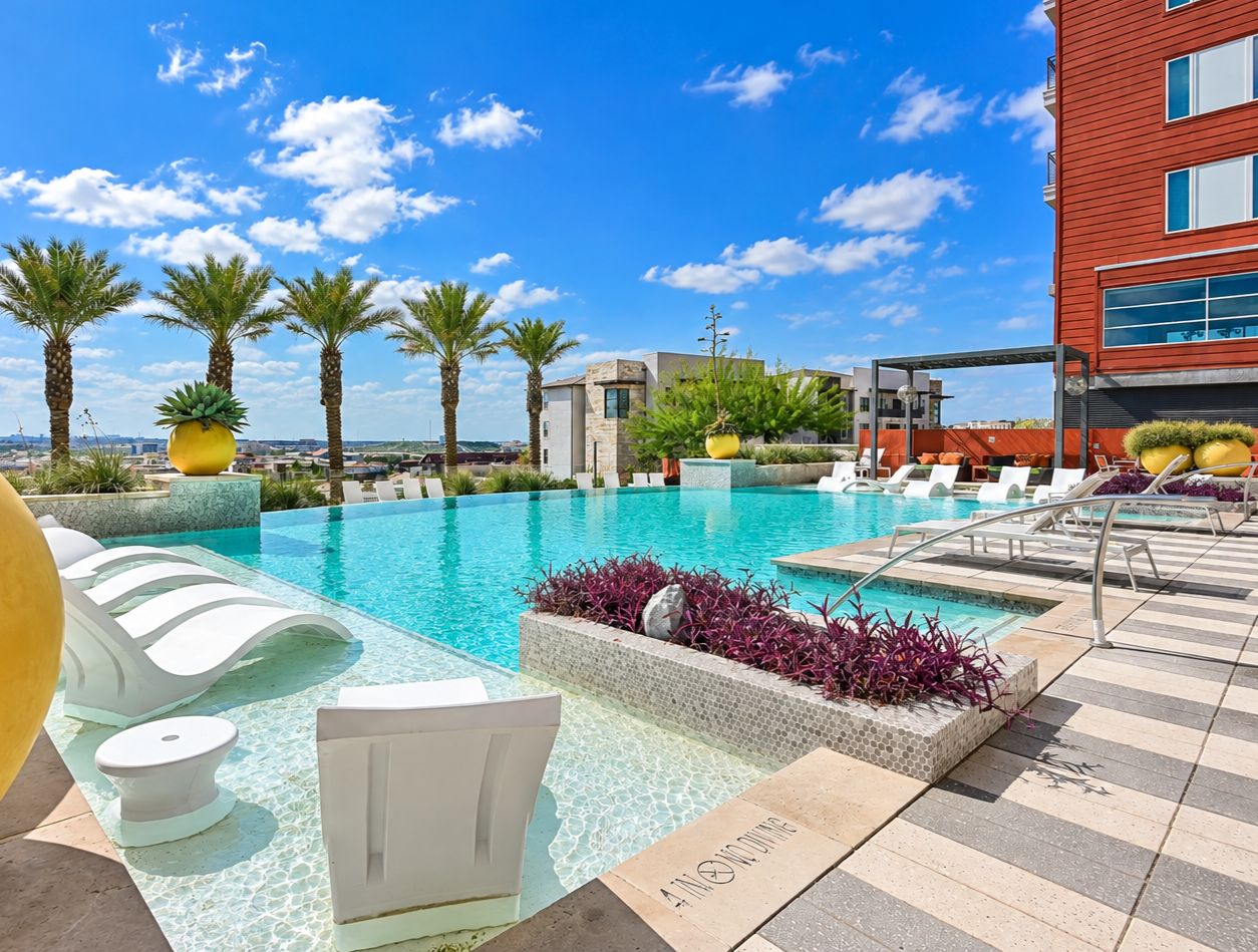 Luxury rooftop swimming pool with lounge chairs, palm trees, vibrant potted plants, and a cityscape view under a partly cloudy blue sky.