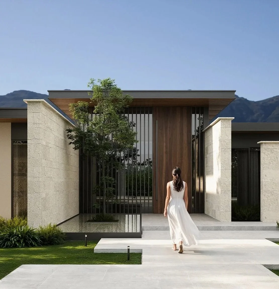 A woman in a white dress walking towards the entrance of a modern house with a large wooden door, stone walls, and a small garden.