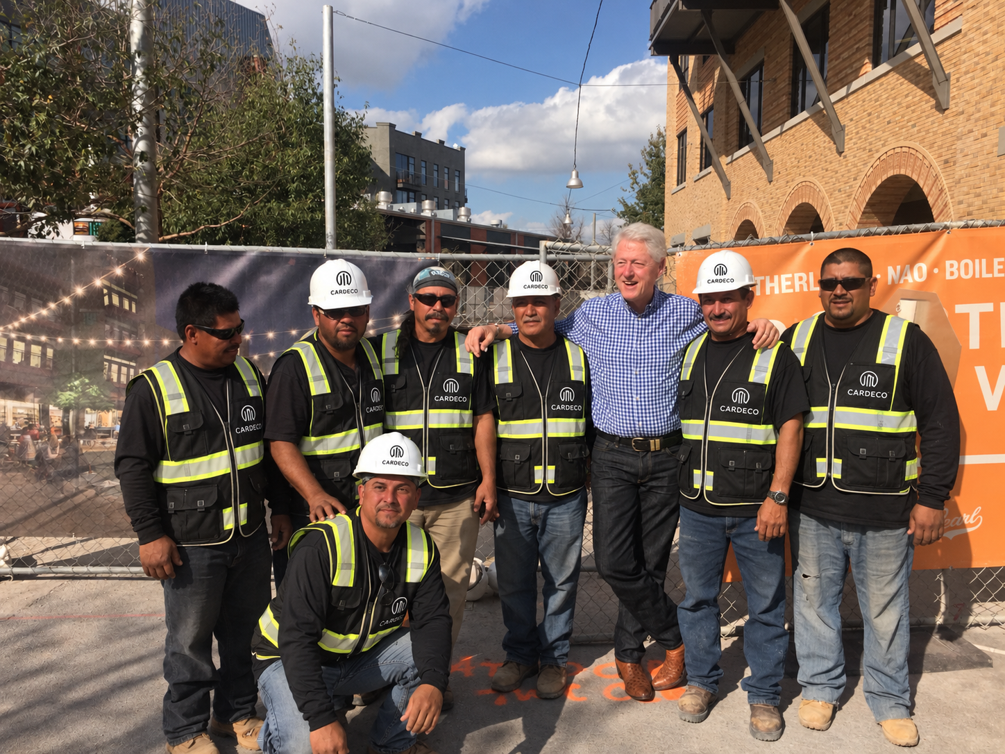 Group of construction workers and a man in blue checkered shirt standing together outdoors at a construction site, some wearing hard hats and safety vests, smiling at the camera.