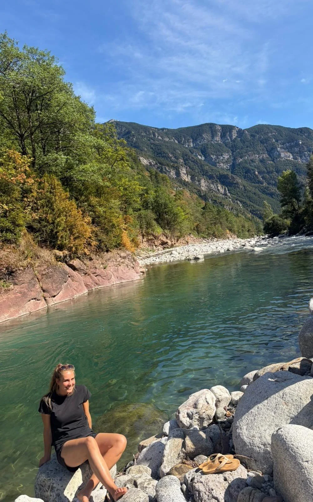 Een vrouw zit op een grote rots naast een rivier, omgeven door stenen en bomen, met bergen en een blauwe hemel op de achtergrond.