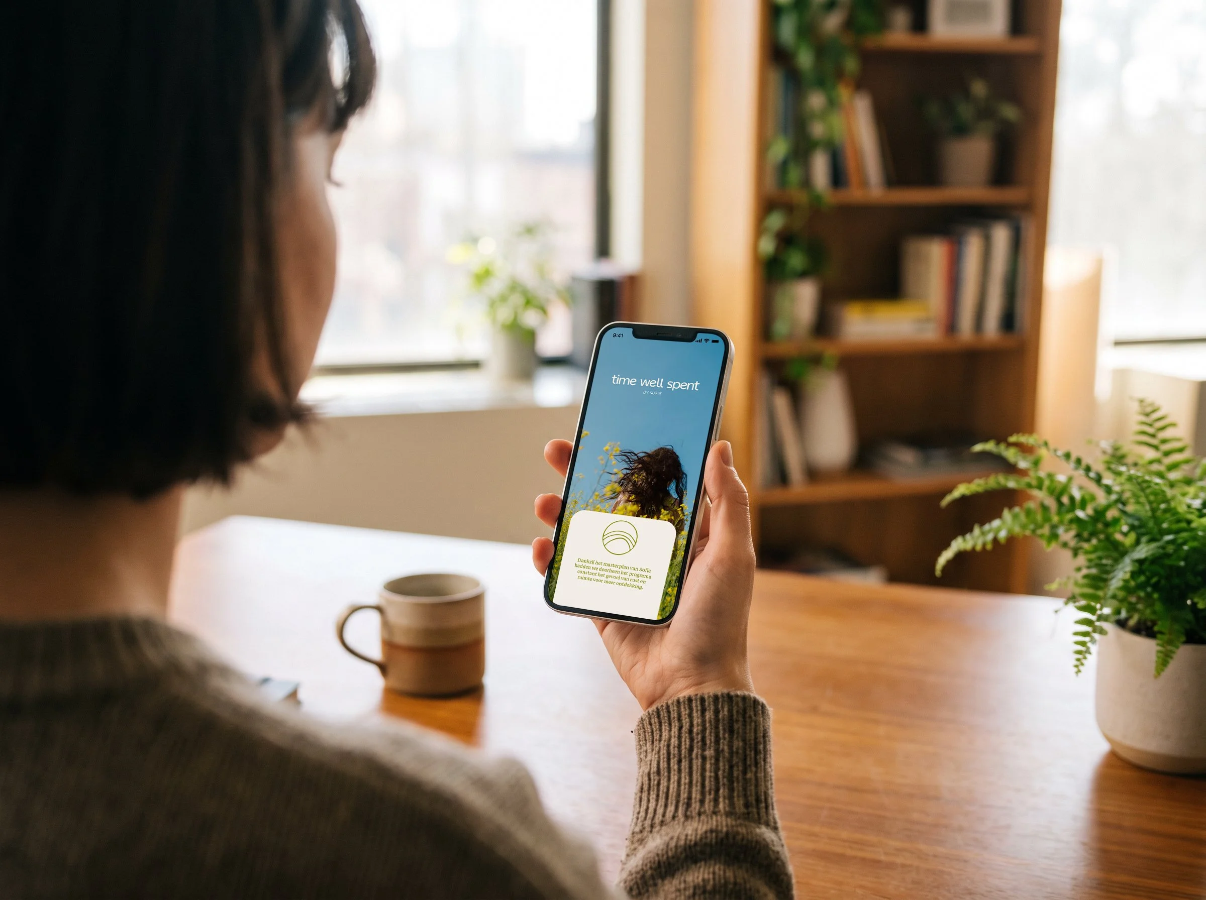 Persoon houdt een smartphone vast die een app toont met de tekst 'time well spent' en een afbeelding van een persoon met lang, golvend haar. Op de achtergrond staan groene planten en een boekenkast, binnen in een huis met veel natuurlijk licht.