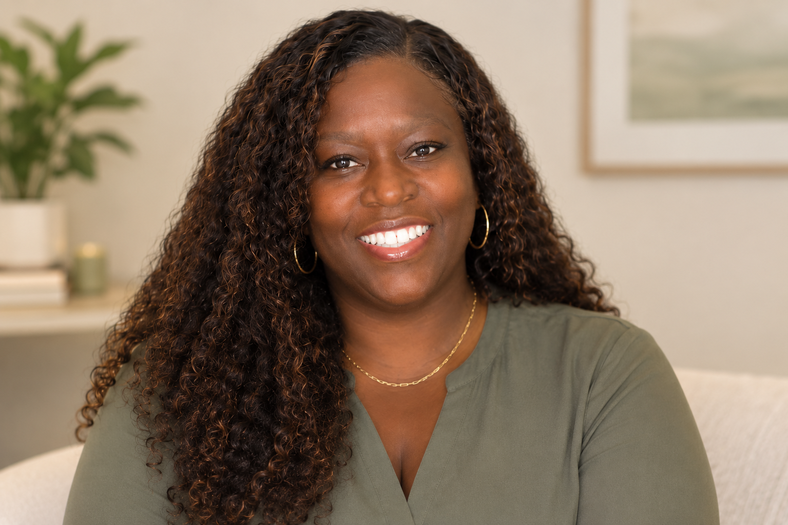A smiling woman with long curly hair wearing gold hoop earrings and a gold necklace, sitting indoors against a neutral background.