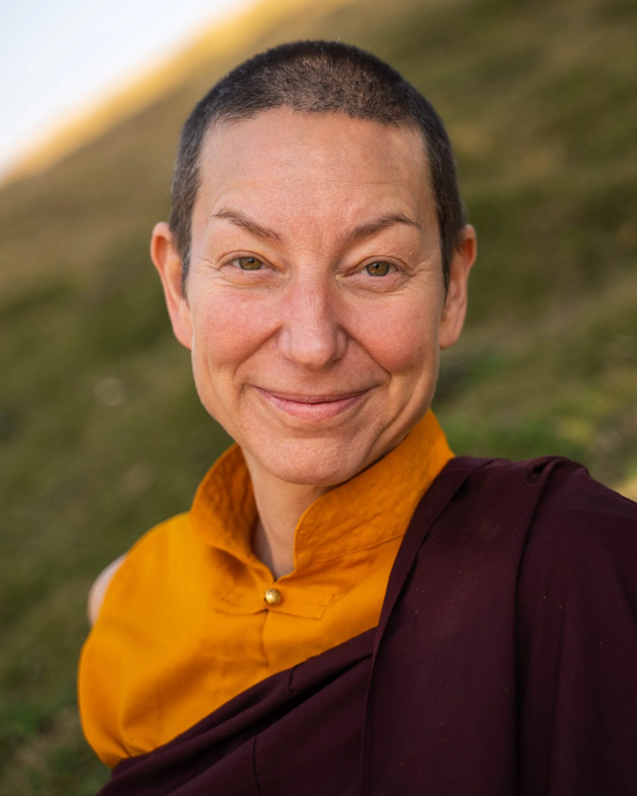 Close-up of a smiling person with short dark hair wearing a maroon and orange outfit, outdoors with blurred greenery in the background.