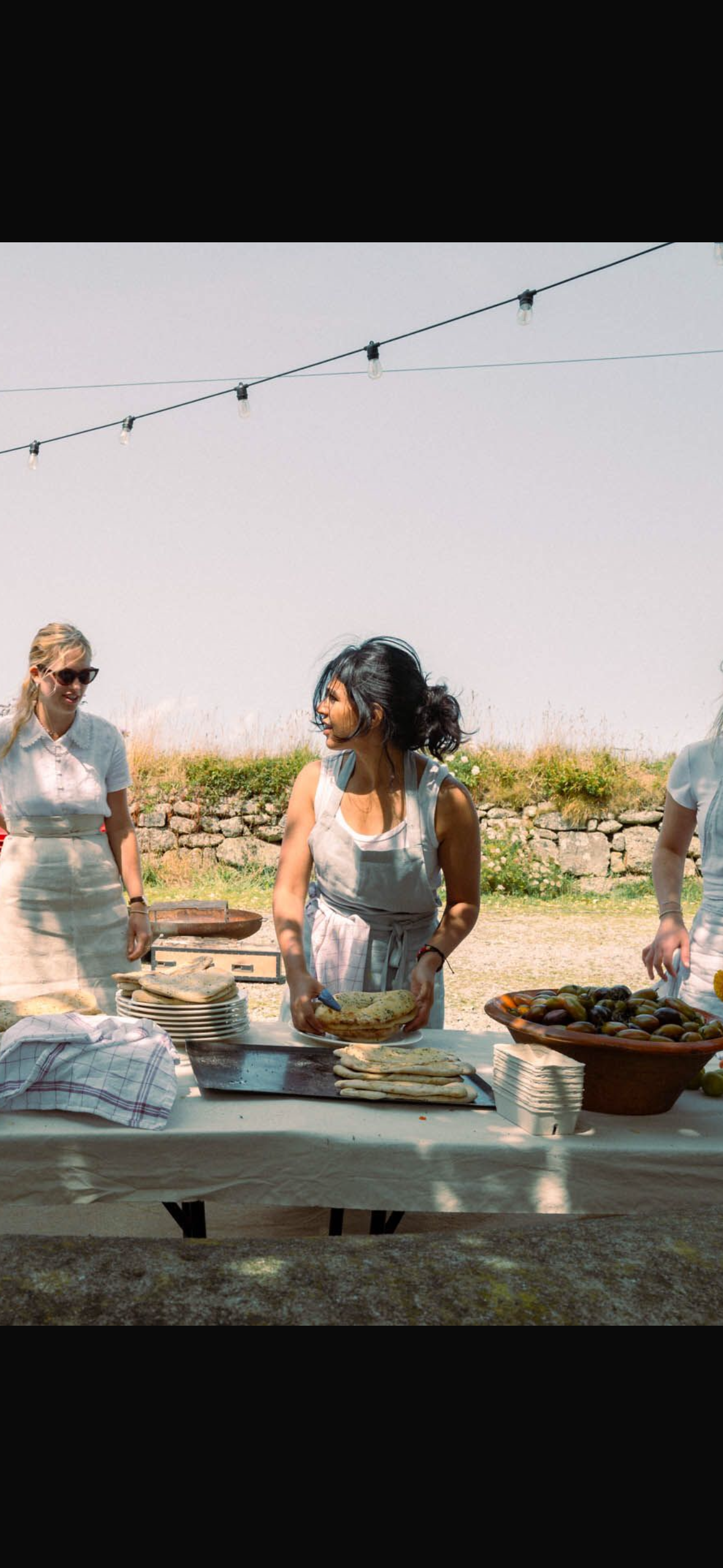 Women at an outdoor food table preparing naan bread, with a stone wall and blue sky in the background.