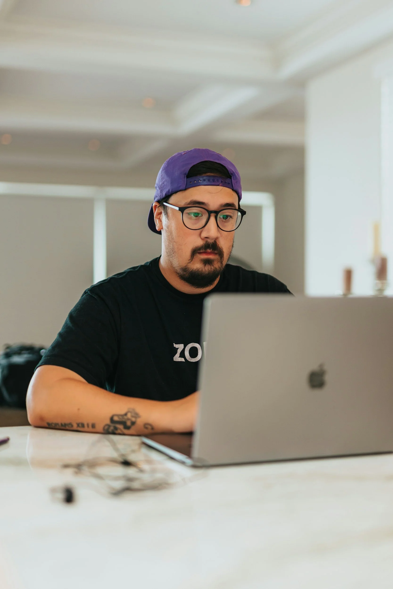 A man wearing glasses, a purple backward baseball cap, and a black t-shirt with white text, sitting at a desk working on a silver MacBook. The man has a beard and tattoos on his left forearm. The background features a bright, minimalistic room with white walls and ceiling, and some blurred objects and furniture.