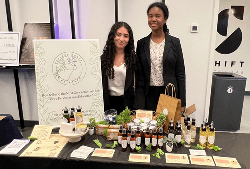 Two women standing behind a table displaying skincare products at an exhibition or event.