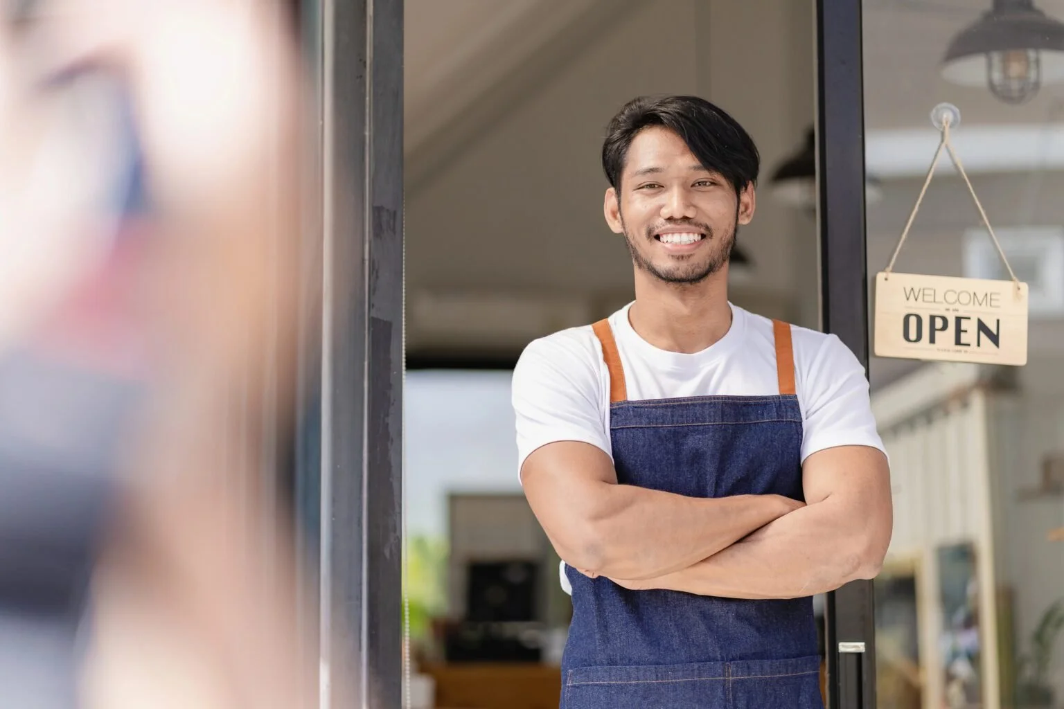 Smiling man in white T-shirt and apron with crossed arms standing at cafe door with 'Welcome Open' sign