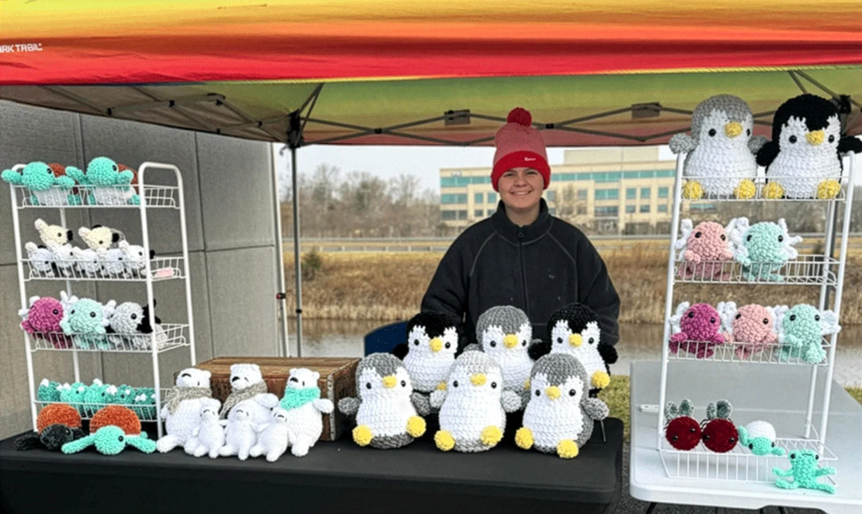 A person standing behind a table of crocheted stuffed penguins and other animals at an outdoor market stall under a red canopy. The table displays various colorful crochet toys shaped like penguins, seals, and crabs. Shelves on either side hold more crocheted animals.