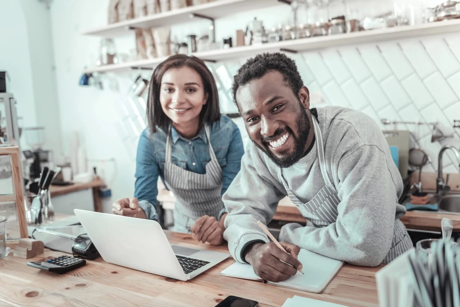 Two smiling people in aprons working in a café or kitchen, one writing in a notebook and the other looking at a laptop on a wooden countertop.