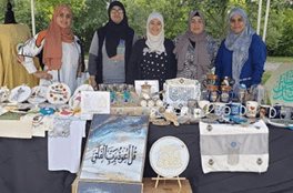 Five women standing behind a table displaying handmade crafts, including pottery, textiles, and decorative items, outdoors with trees in the background.