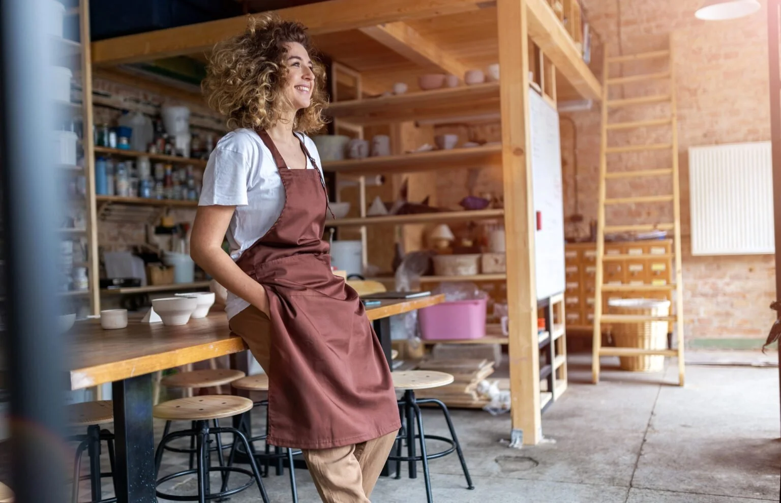 A woman with curly hair wearing a white t-shirt and a brown apron, sitting on a stool in a pottery studio, smiling with her hands in her apron pockets.