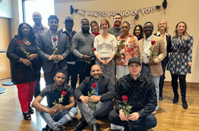 Group of diverse people posing indoors, some holding red roses, in front of a banner that reads 'We Are So Proud of You!'
